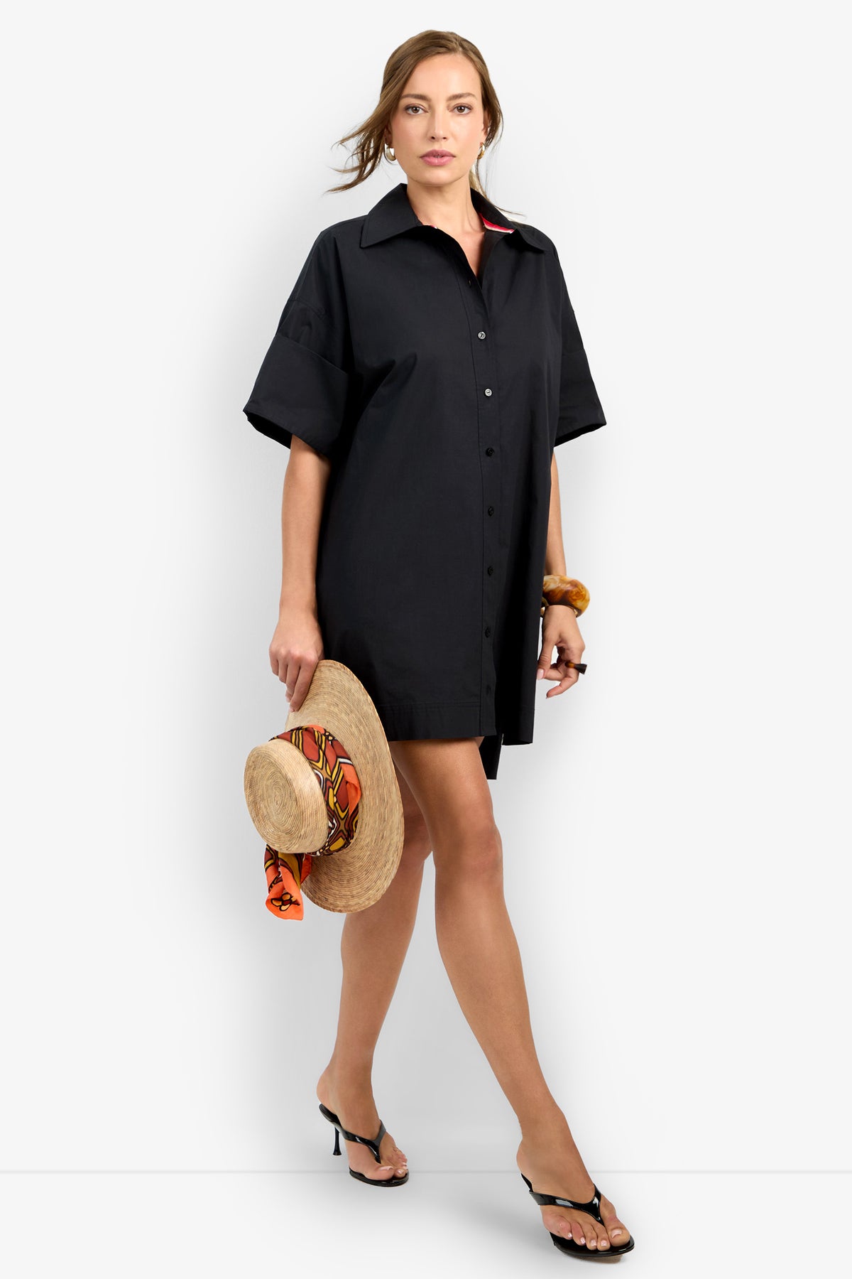 A woman wearing the loose-fitting black OPPONENT DRESS and black heeled sandals holds a straw hat with an orange scarf while walking forward against a plain white background.