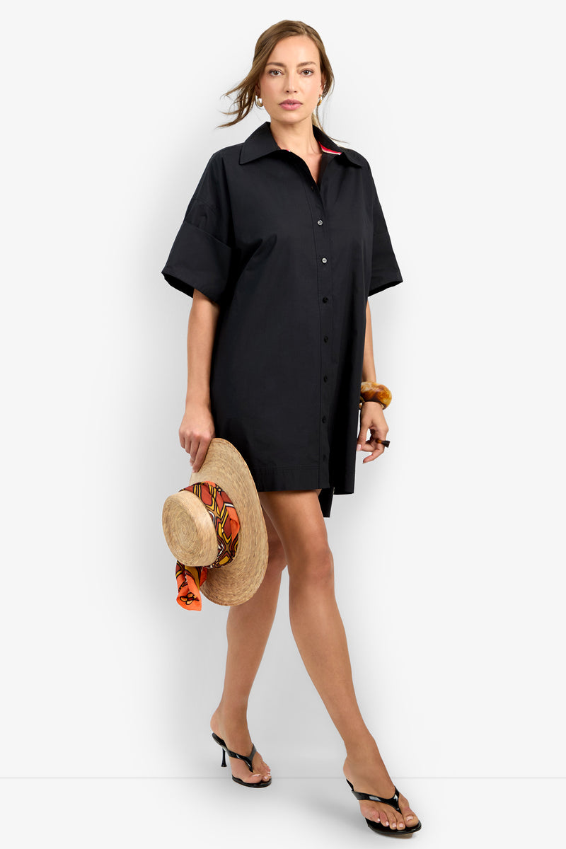 A woman wearing the loose-fitting black OPPONENT DRESS and black heeled sandals holds a straw hat with an orange scarf while walking forward against a plain white background.