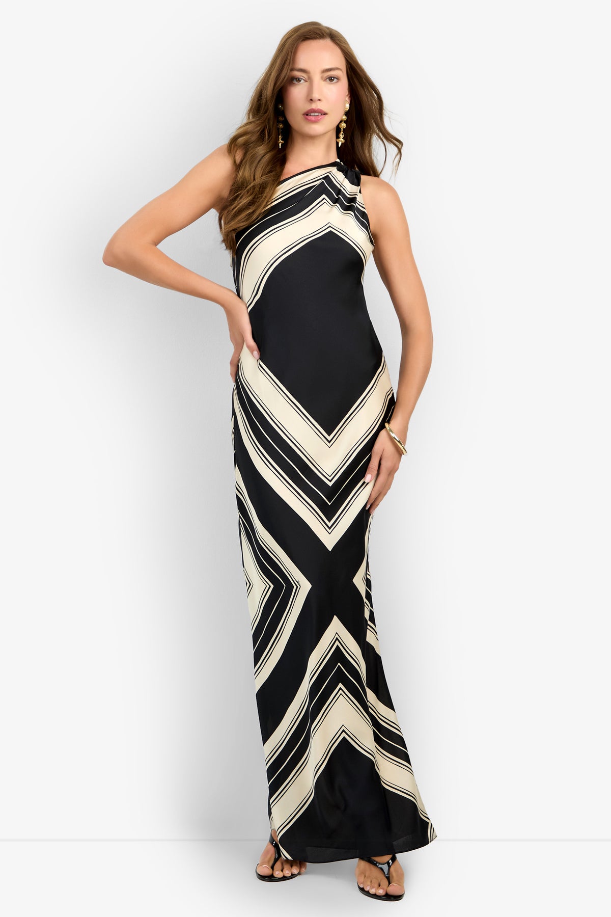 A person poses in the CHIANTI DRESS, a one-shoulder, floor-length black gown with bold cream geometric stripes, paired with sandals and earrings, against a plain white background.