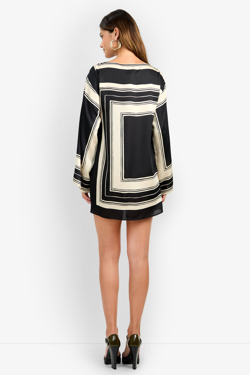 A woman with long brown hair and large hoop earrings faces away, wearing the YULIAH DRESS—a long-sleeve, loose black and cream geometric-patterned mini dress with sheer sleeves—paired with black high heels.