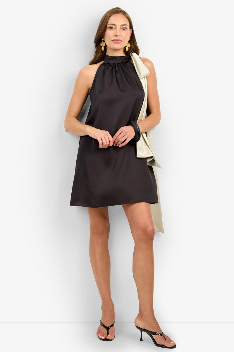 Wearing the VANNI DRESS—a sleeveless, knee-length black dress with a high neckline and off-white draped shoulder detail—a woman pairs it with gold earrings, black bangles, and black heeled sandals.