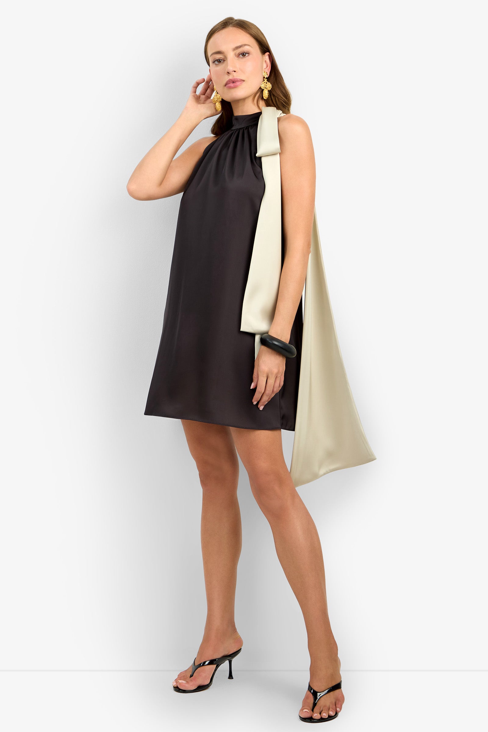 A woman poses in the VANNI DRESS, a sleeveless black mini dress with a gathered neckline and an oversized cream bow on one shoulder, paired with black heeled sandals, gold earrings, and a black bangle.