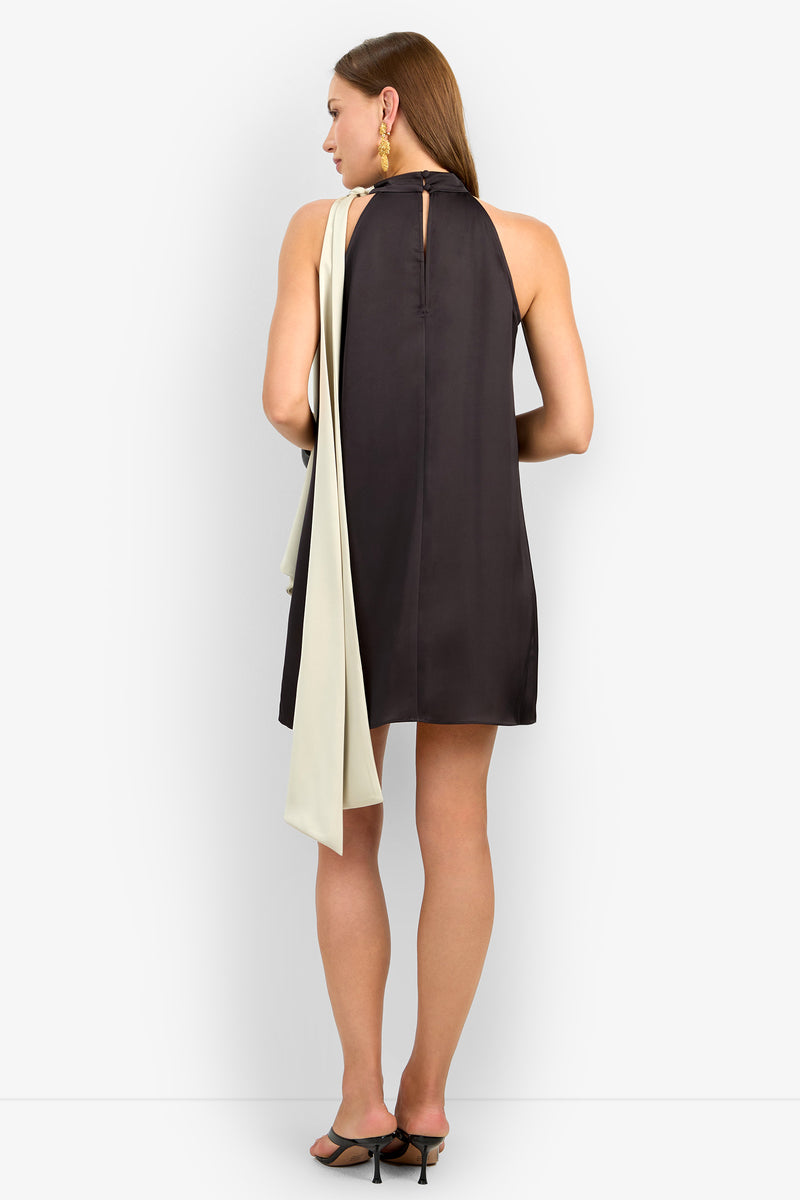 A woman with straight hair, gold earrings, and black heeled sandals wears the VANNI DRESS: a sleeveless short black dress featuring a keyhole back and a long cream drape over one shoulder, standing facing away.