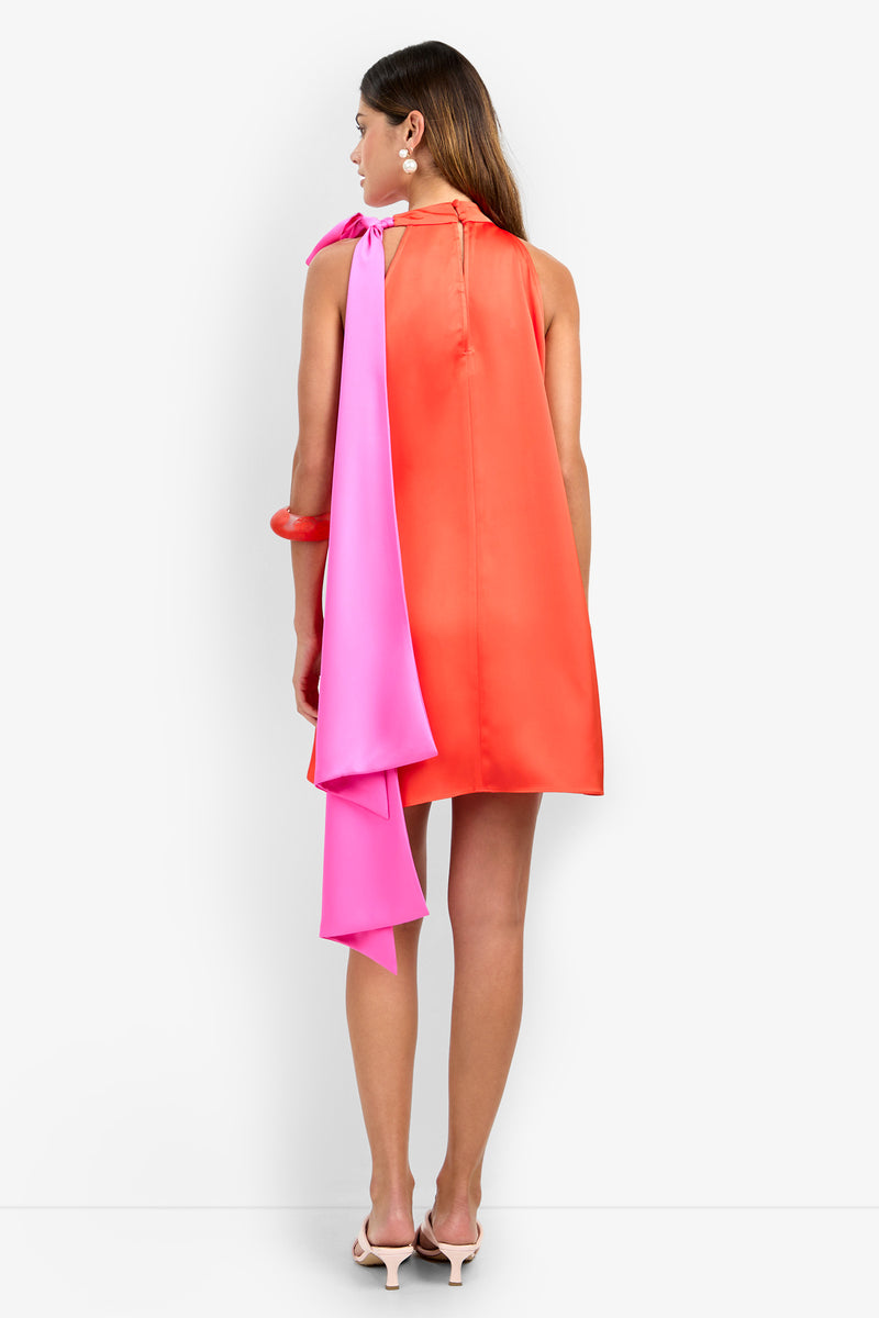 A woman with long brown hair stands facing away on a plain white background, wearing the sleeveless, loose-fitting VANNI DRESS in red, paired with a long pink scarf over one shoulder, white earrings, and white heels.