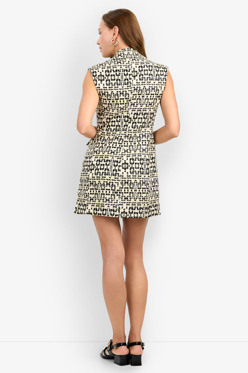 A woman with straight light brown hair stands facing away, wearing the BAOBAB DRESS—a sleeveless dress with black and beige geometric patterns. She pairs it with black sandals and hoop earrings against a plain white background.