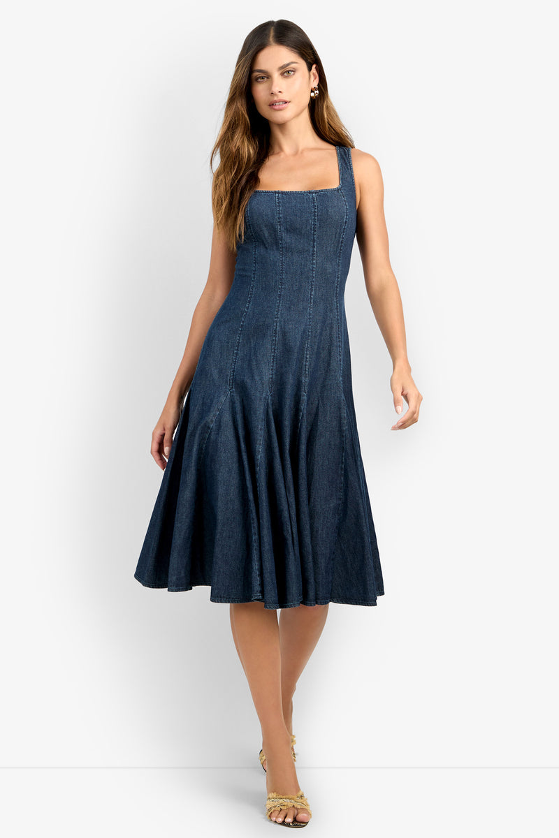 A woman wearing the CASIMA DRESS, a sleeveless, square-neck dark blue denim midi with a flared skirt, and beige heeled sandals stands against a white background.