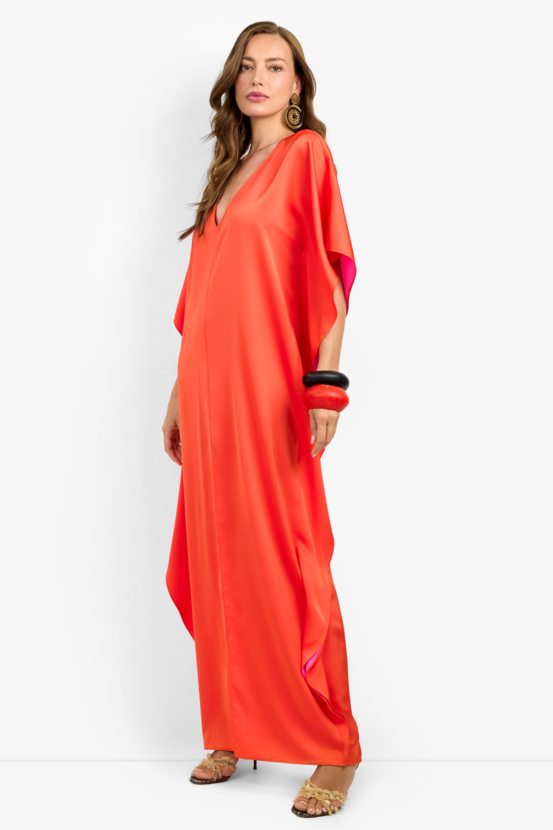 A woman poses against a white background wearing the RIVA DRESS— a loose, floor-length, bright orange satin dress with short sleeves. She styles it with long wavy hair, hoop earrings, chunky bangles, and strappy heels.