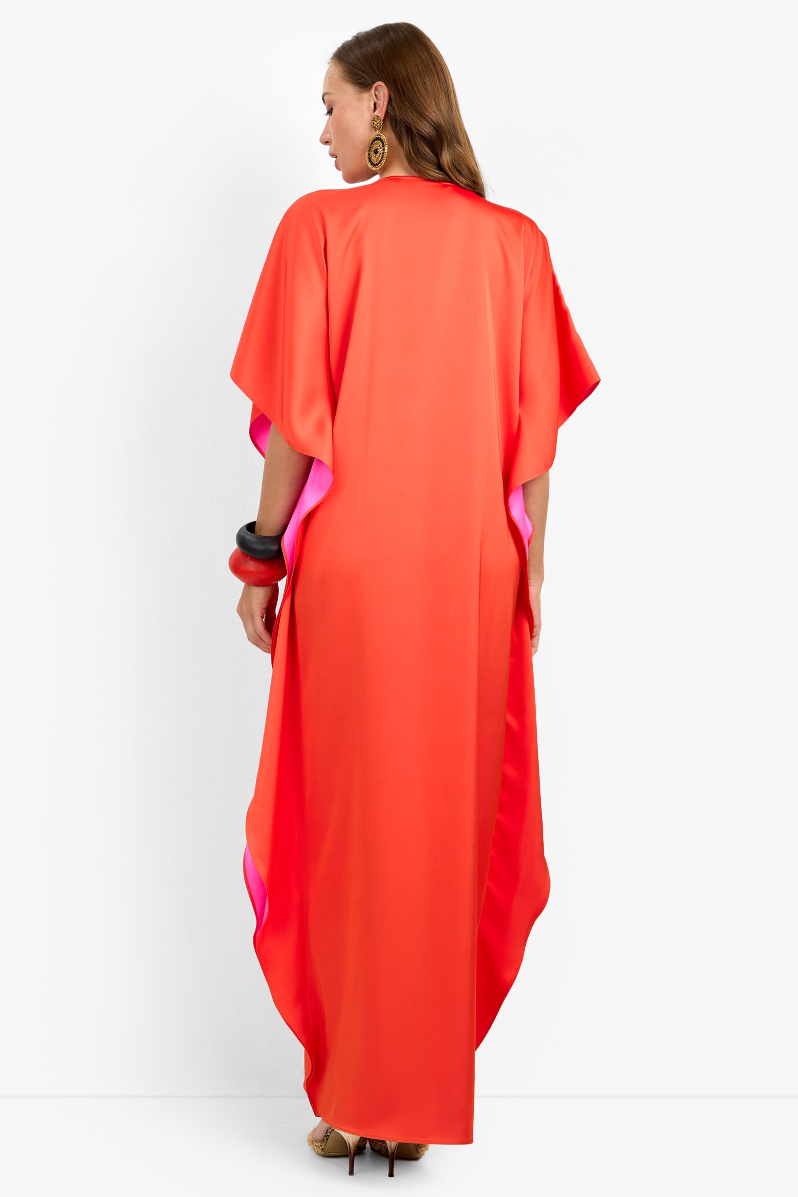 A woman stands facing away, wearing the RIVA DRESS—a long, loose-fitting, bright red and pink kaftan with short sleeves. She accessorizes with large gold earrings, chunky bangles, and beige high heels.