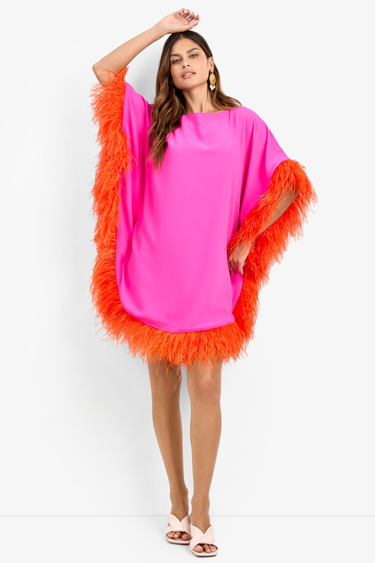 A woman models the MINI NEENA, a loose, hot pink dress with bright orange feather trim on the sleeves and hem. She pairs it with white heeled sandals and gold earrings, her hair in loose waves and one arm raised.
