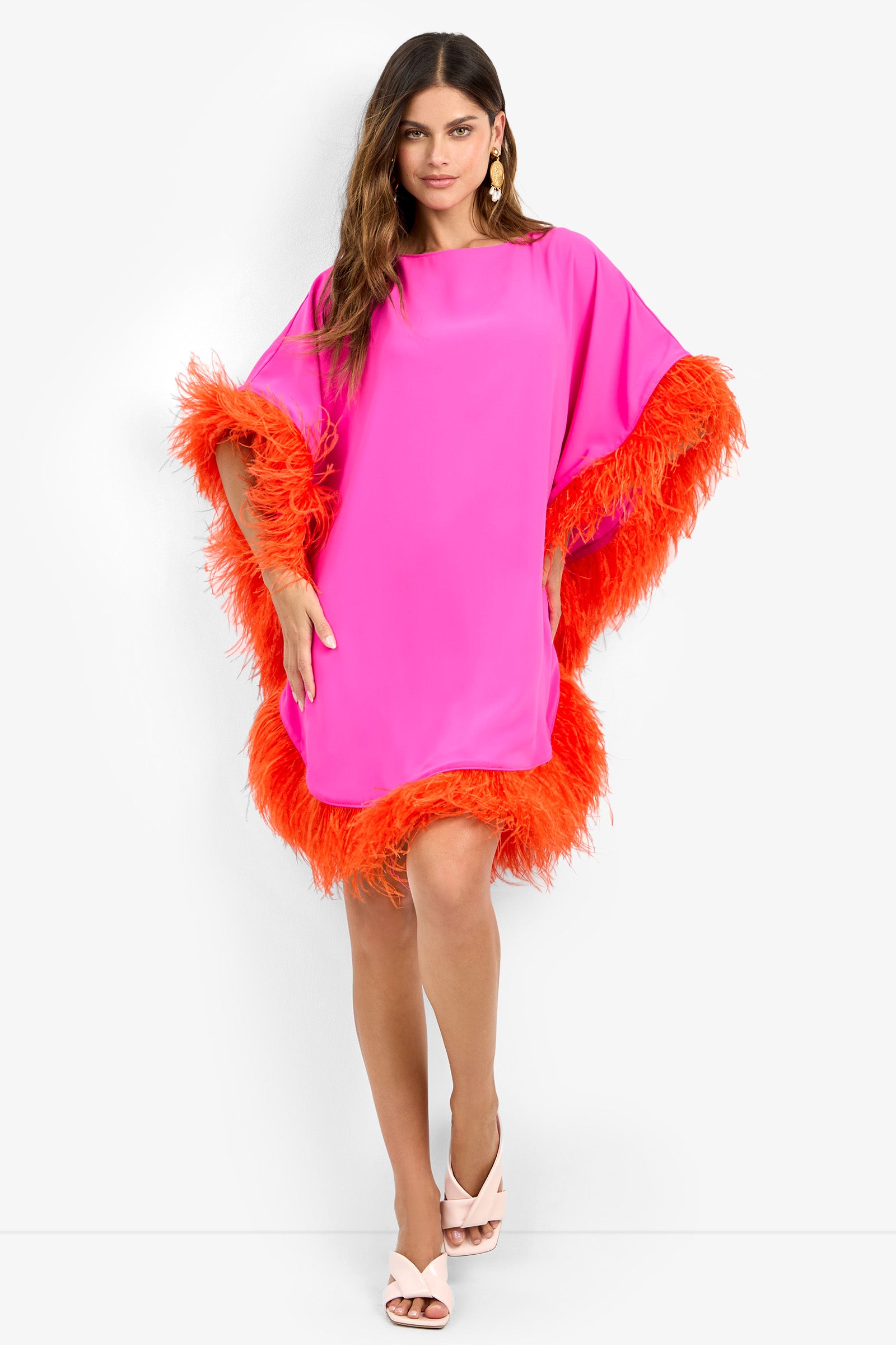 Woman in the MINI NEENA, a loose-fitting bright pink dress with orange feather trim on sleeves and hem, poses against a white background wearing light pink heels and statement earrings.