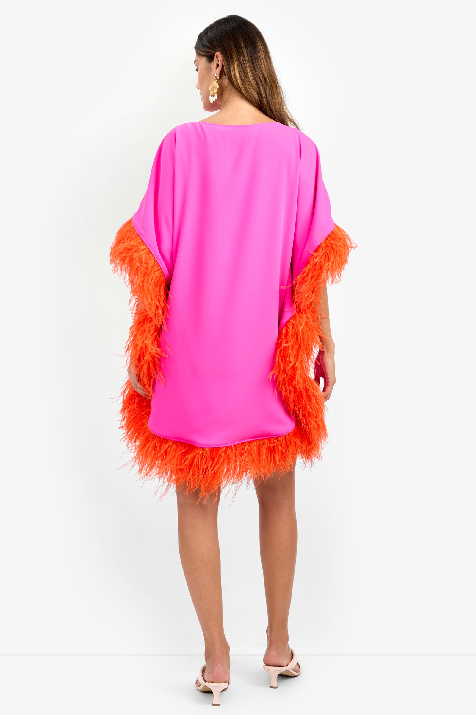 A woman faces away, wearing the MINI NEENA—a loose, bright pink dress with orange feather trim on the sleeves and hem. She has long brown hair, earrings, and white heeled sandals.