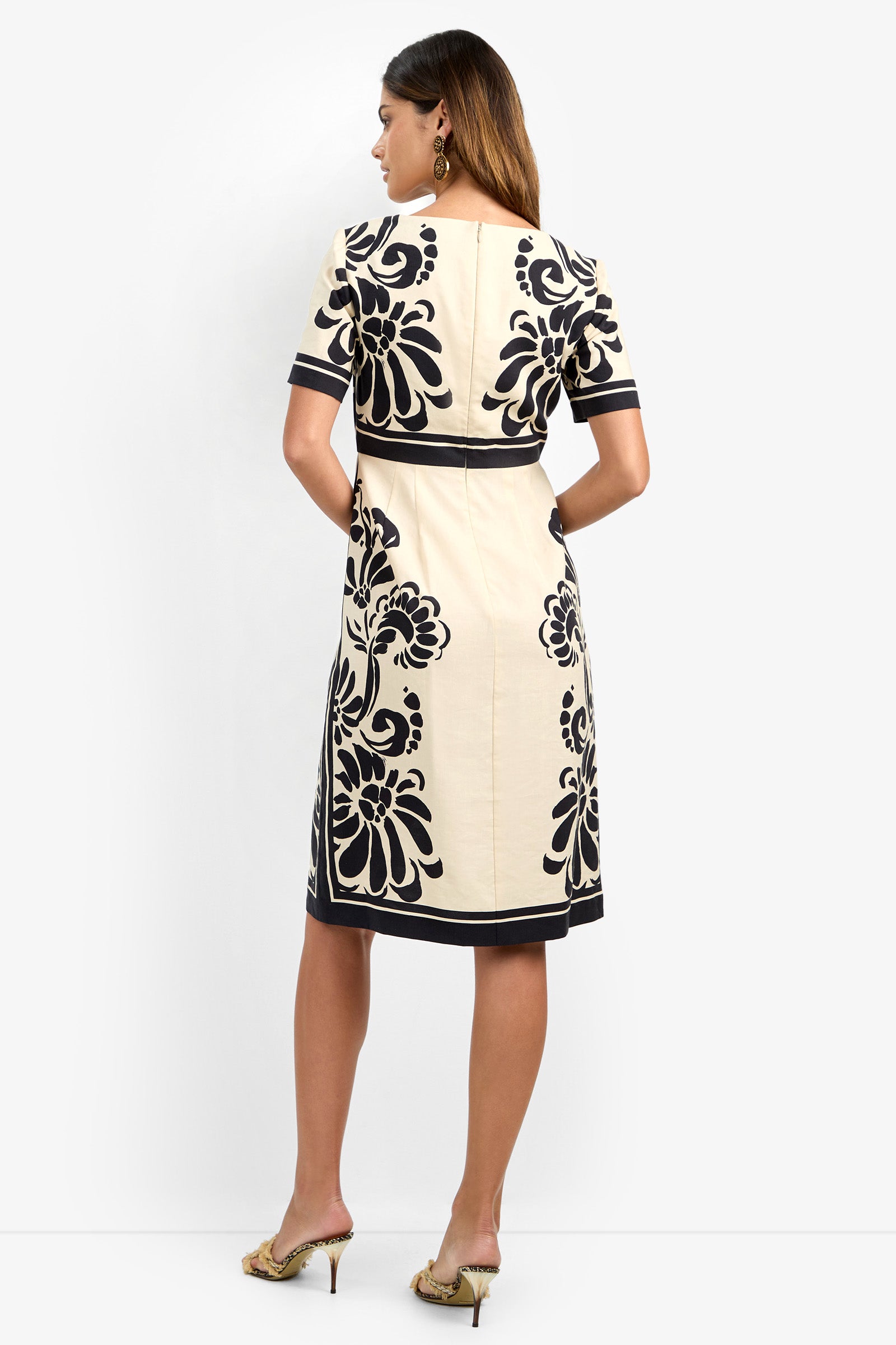 A woman faces away, modeling the JENNAH DRESS—a knee-length cream dress with bold black floral patterns, short sleeves, and black edging. She pairs it with heels and dangling earrings, hands on her hips.