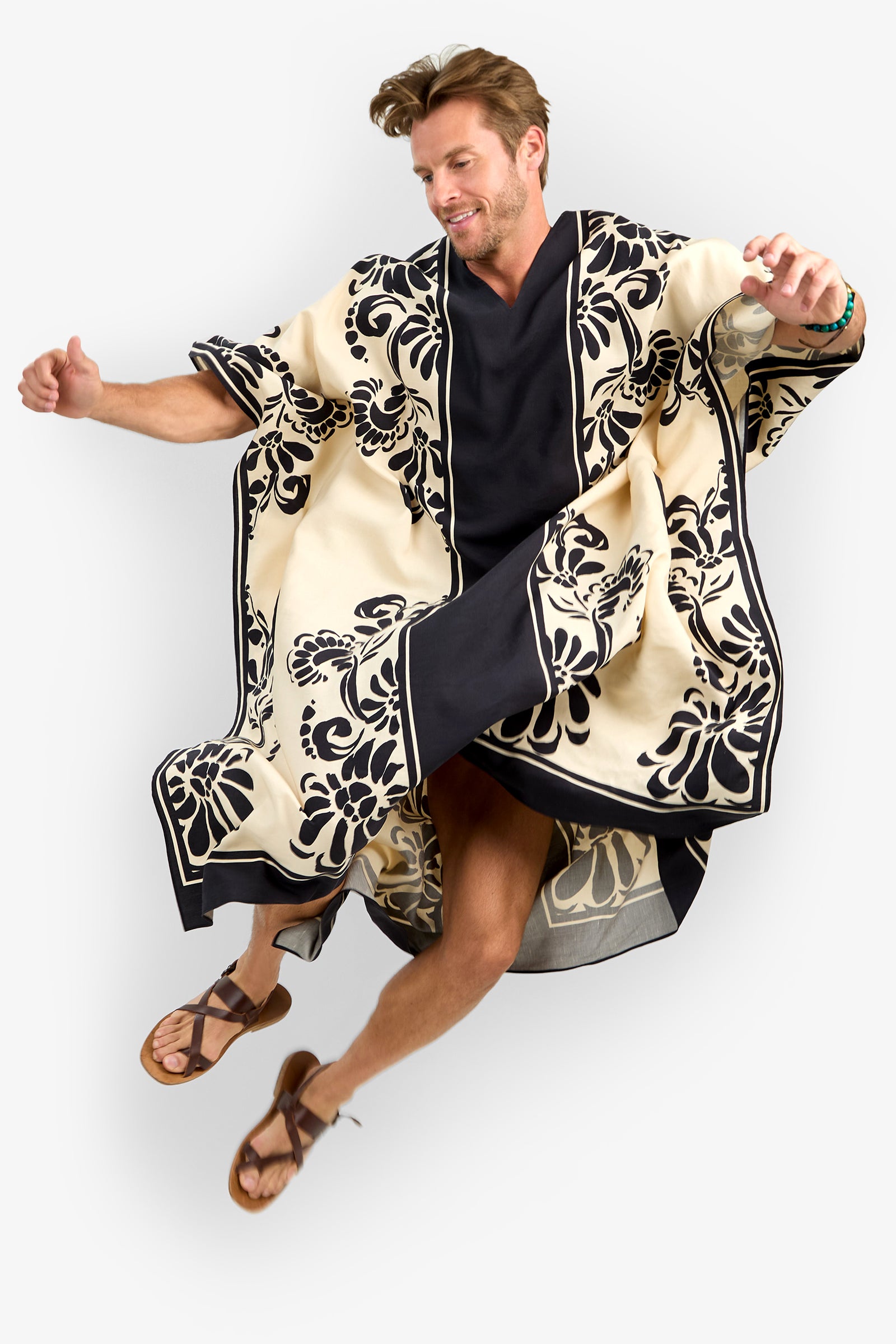 A man wearing the SAN MARTINO CAFTAN, a cream and black floral patterned robe with wide sleeves, smiles while jumping in brown sandals and a black shirt against a white background.