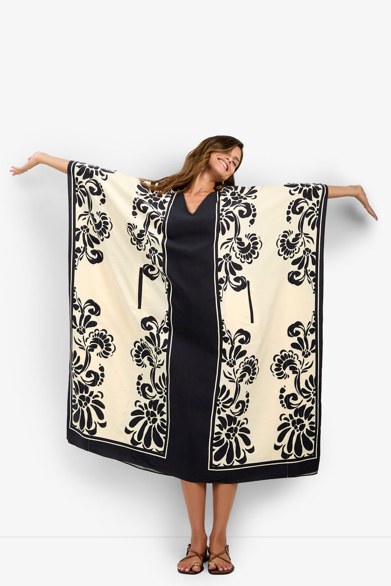 A woman stands with arms outstretched against a plain white background, wearing the San Martino Caftan—a cream and black floral-patterned kimono-style robe—over a black dress and sandals.