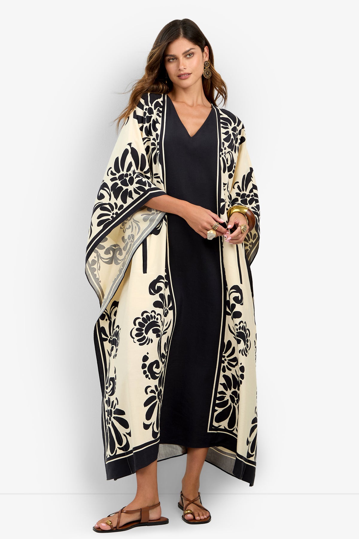 A woman wearing the San Martino Caftan—a long, cream-colored kimono adorned with bold black floral designs—layered over a black dress and paired with sandals, stands against a plain white background.