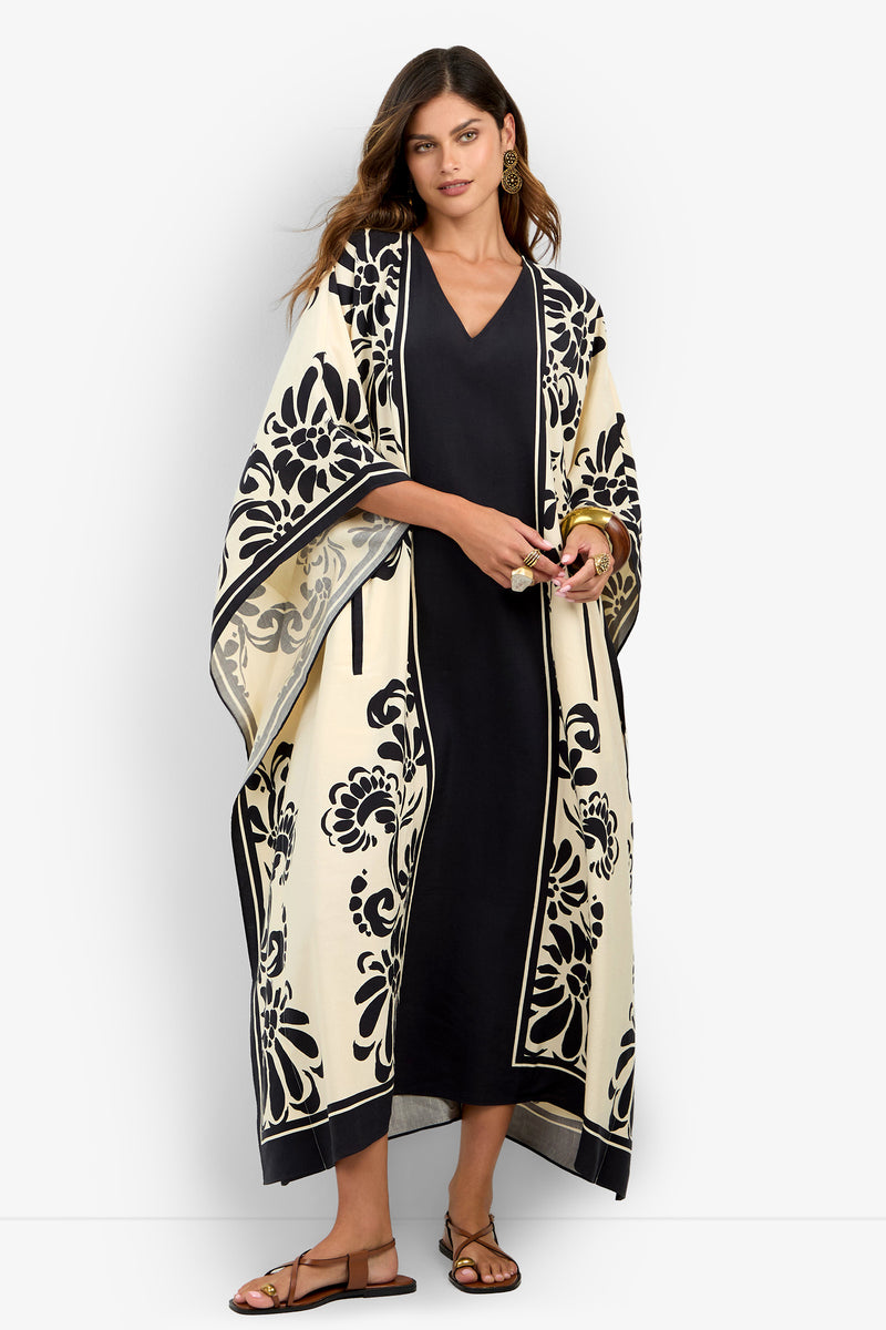 A woman wearing the San Martino Caftan—a long, cream-colored kimono adorned with bold black floral designs—layered over a black dress and paired with sandals, stands against a plain white background.