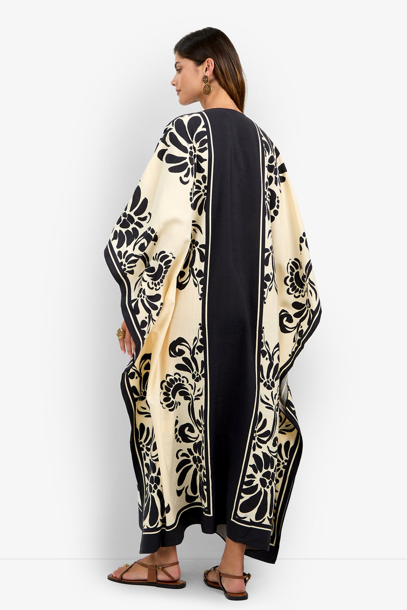 A woman stands sideways against a plain white background, wearing the San Martino Caftan—long and loose with a cream and black floral pattern—paired with black sandals, large earrings, rings, and her hair down.