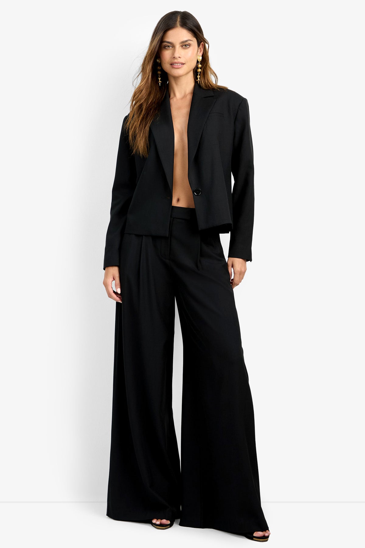 A woman with long brown hair stands against a white background, wearing the MALINE JACKET over bare skin, wide-leg black pants, open-toe black heels, and gold earrings. She faces forward with a neutral expression.