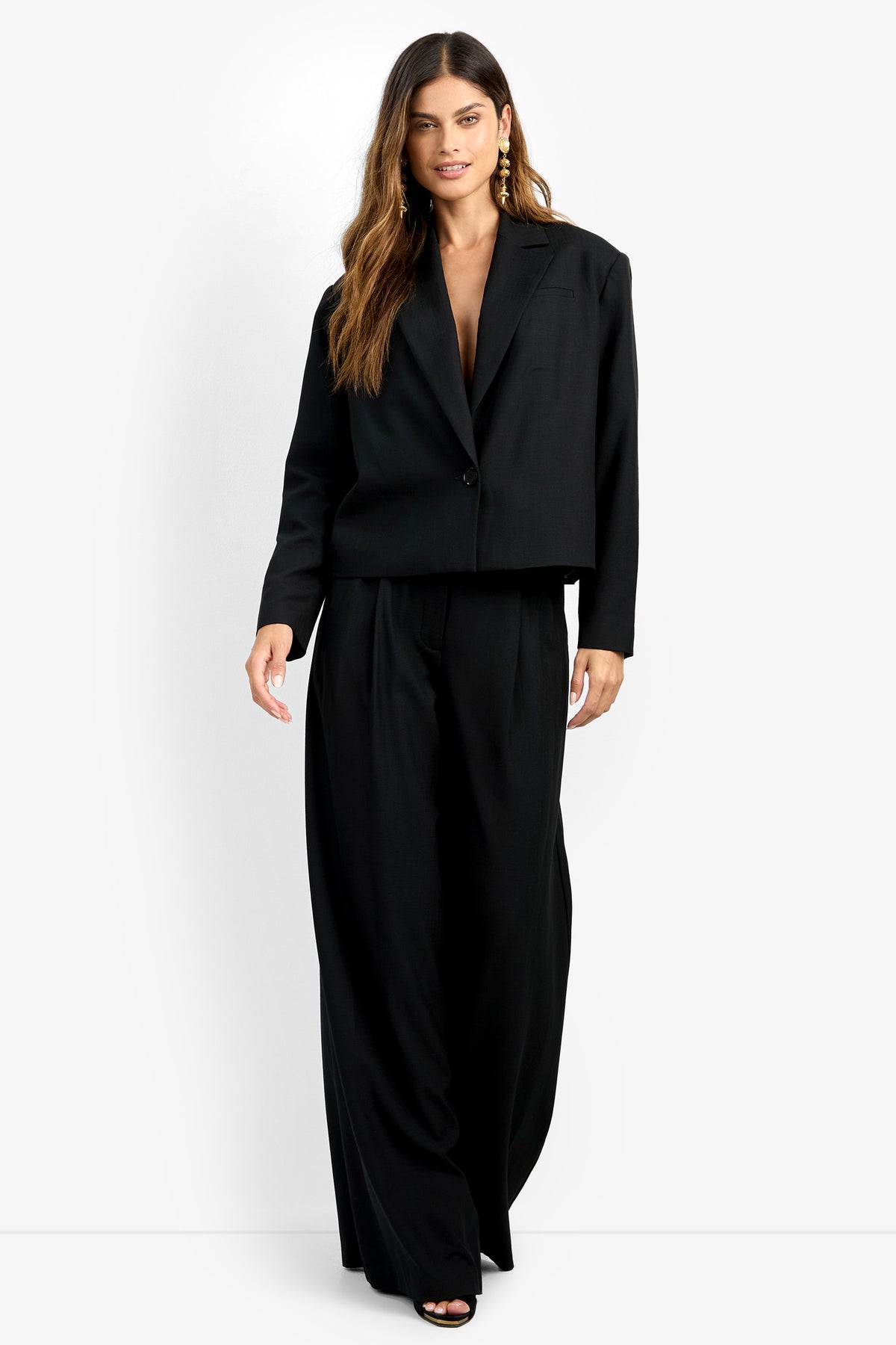 A woman with long brown hair wears the loose-fitting MALINE JACKET and wide-leg black trousers, standing forward against a white background with one hand at her side and the other slightly raised.