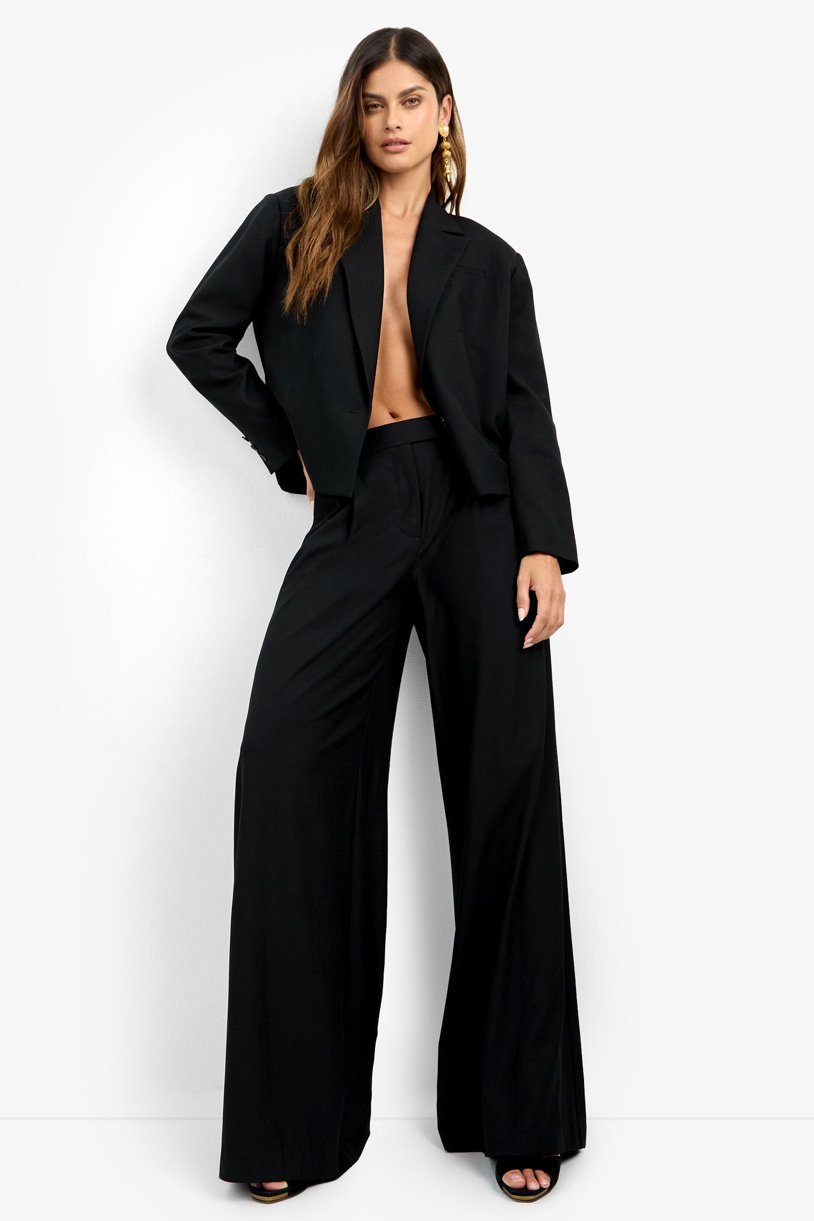 A woman poses confidently against a white background wearing the MIMMO PANT, a loose-fitting black wide-leg trouser. She pairs it with black open-toe heels, gold earrings, and leaves her torso bare beneath the suit jacket.