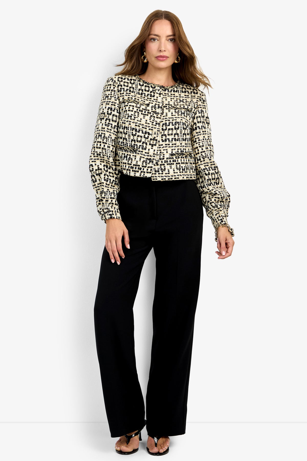 A woman with loose hair and a confident pose stands against a plain background wearing the GIULIA JACKET, styled with black wide-leg pants and black heeled sandals.