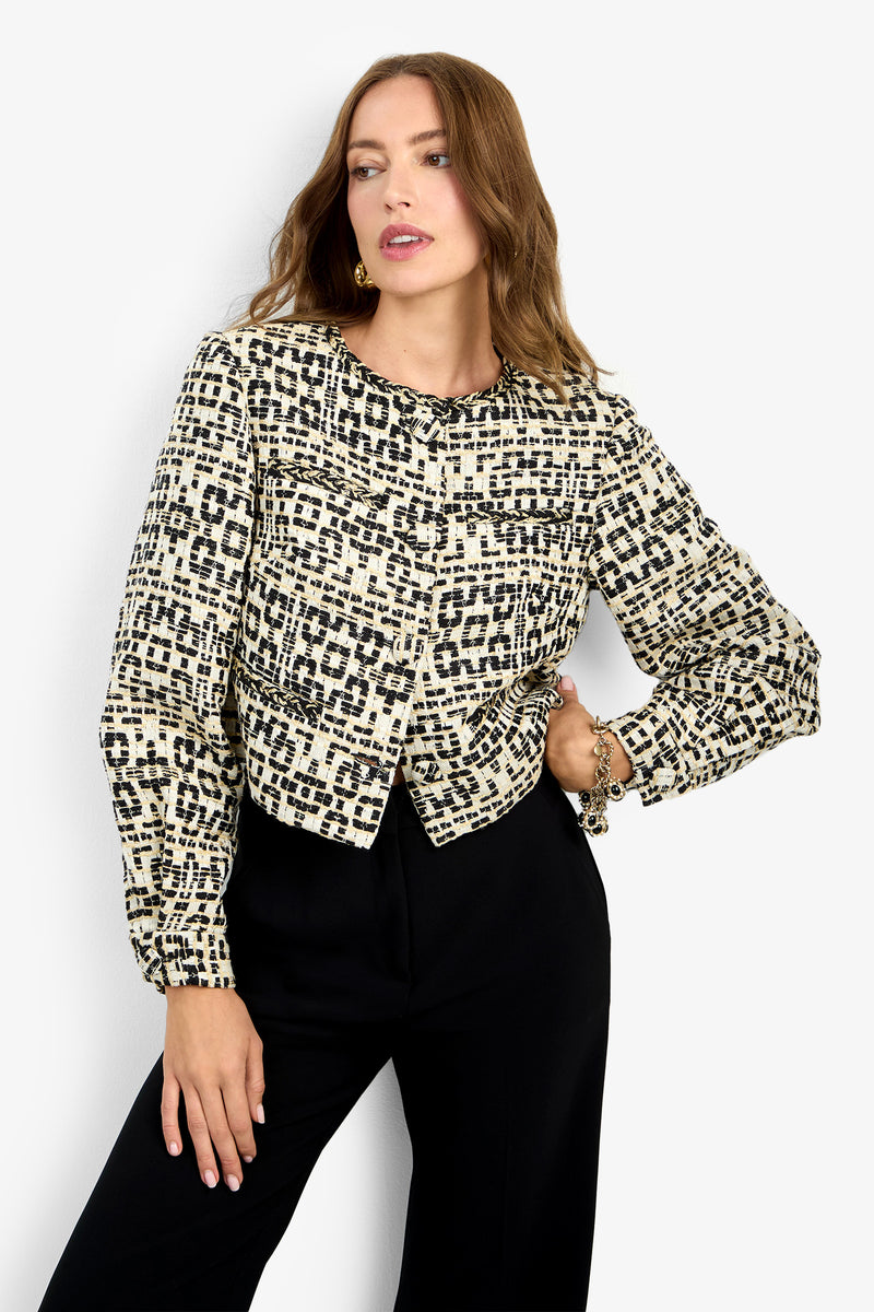A woman with long brown hair wears the GIULIA JACKET—a patterned, cropped, long-sleeve jacket with black and cream geometric designs—paired with black high-waisted pants, posing against a plain light background.