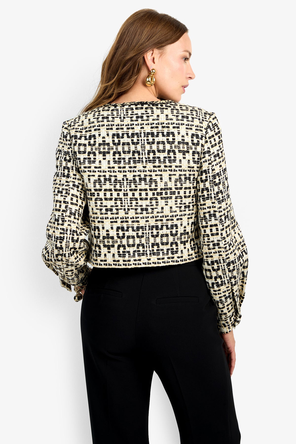 A woman with long brown hair wears the GIULIA JACKET, featuring a black and white geometric pattern and long sleeves, paired with black pants. She is shown from the back against a plain light background.