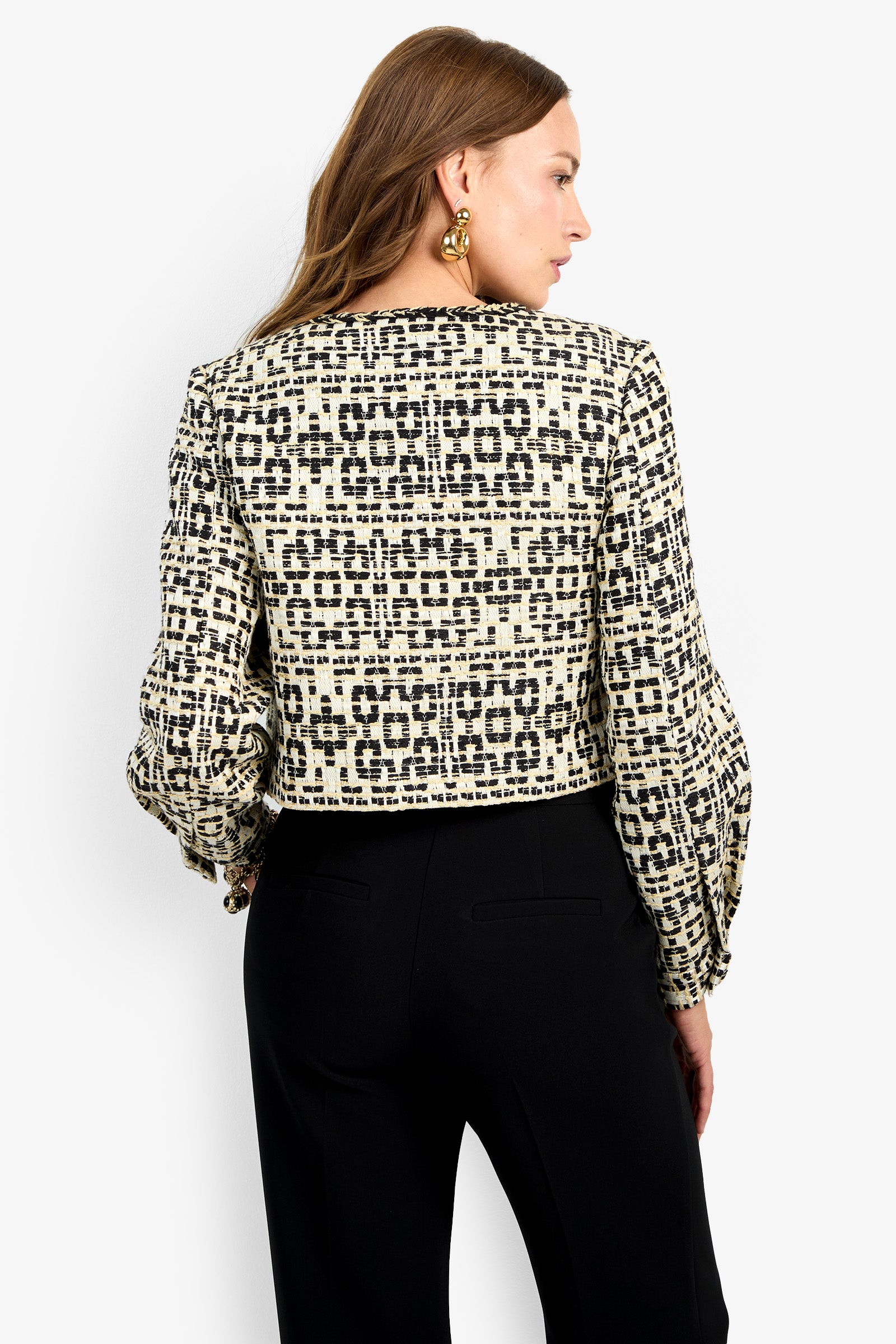 A woman with long brown hair wears the GIULIA JACKET, featuring a black and white geometric pattern and long sleeves, paired with black pants. She is shown from the back against a plain light background.