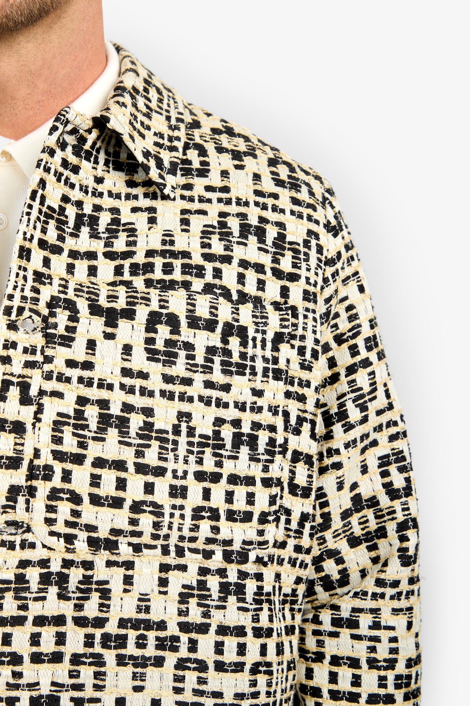 A close-up of someone wearing the PIERRE JACKET with a black and beige geometric texture, a large chest pocket, and a collared neckline. Only part of their face is visible.