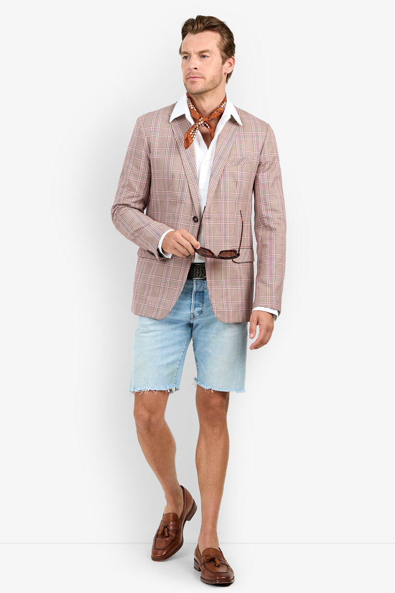 A man wears the THURSTON BLAZER, white shirt, brown scarf, light denim shorts, brown belt, and loafers. Holding sunglasses, he looks to the side against a plain background.