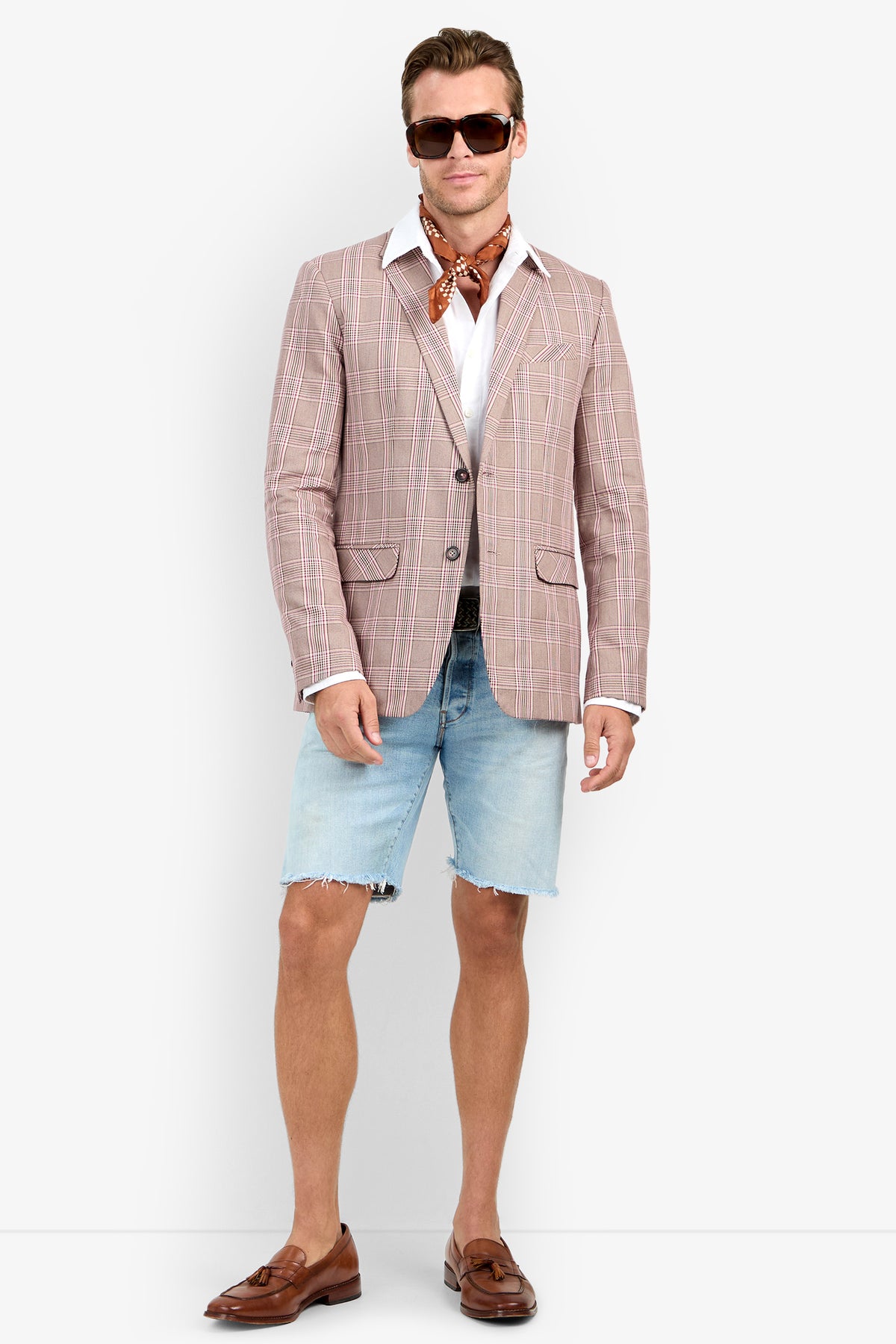 A man wears sunglasses, a white shirt, brown scarf, blue denim shorts, brown loafers, and the THURSTON BLAZER in light brown plaid, standing against a plain background.