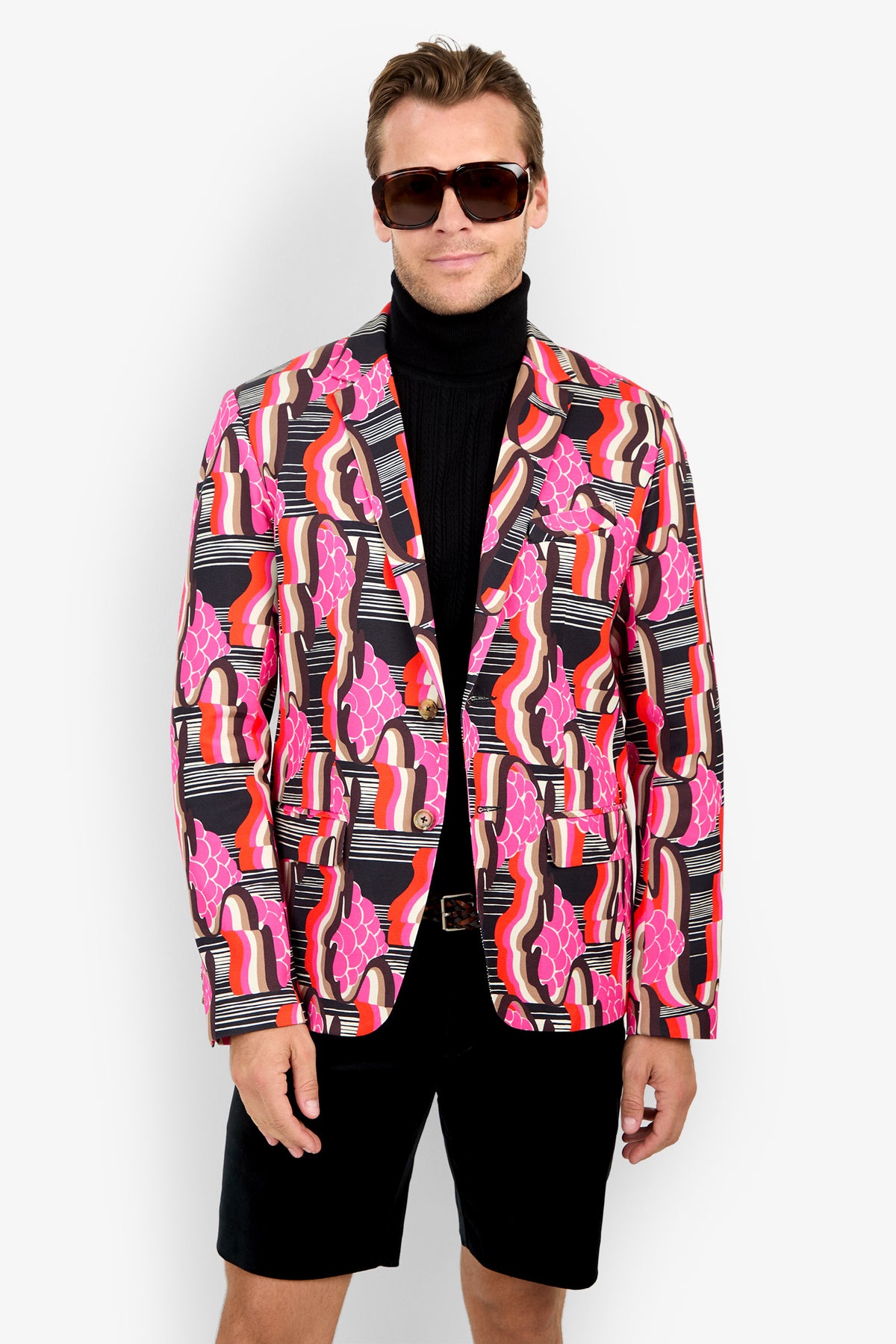 A man wearing sunglasses, a black turtleneck, black shorts, and the THURSTON BLAZER featuring pink, red, white, and black patterns stands against a plain white background.