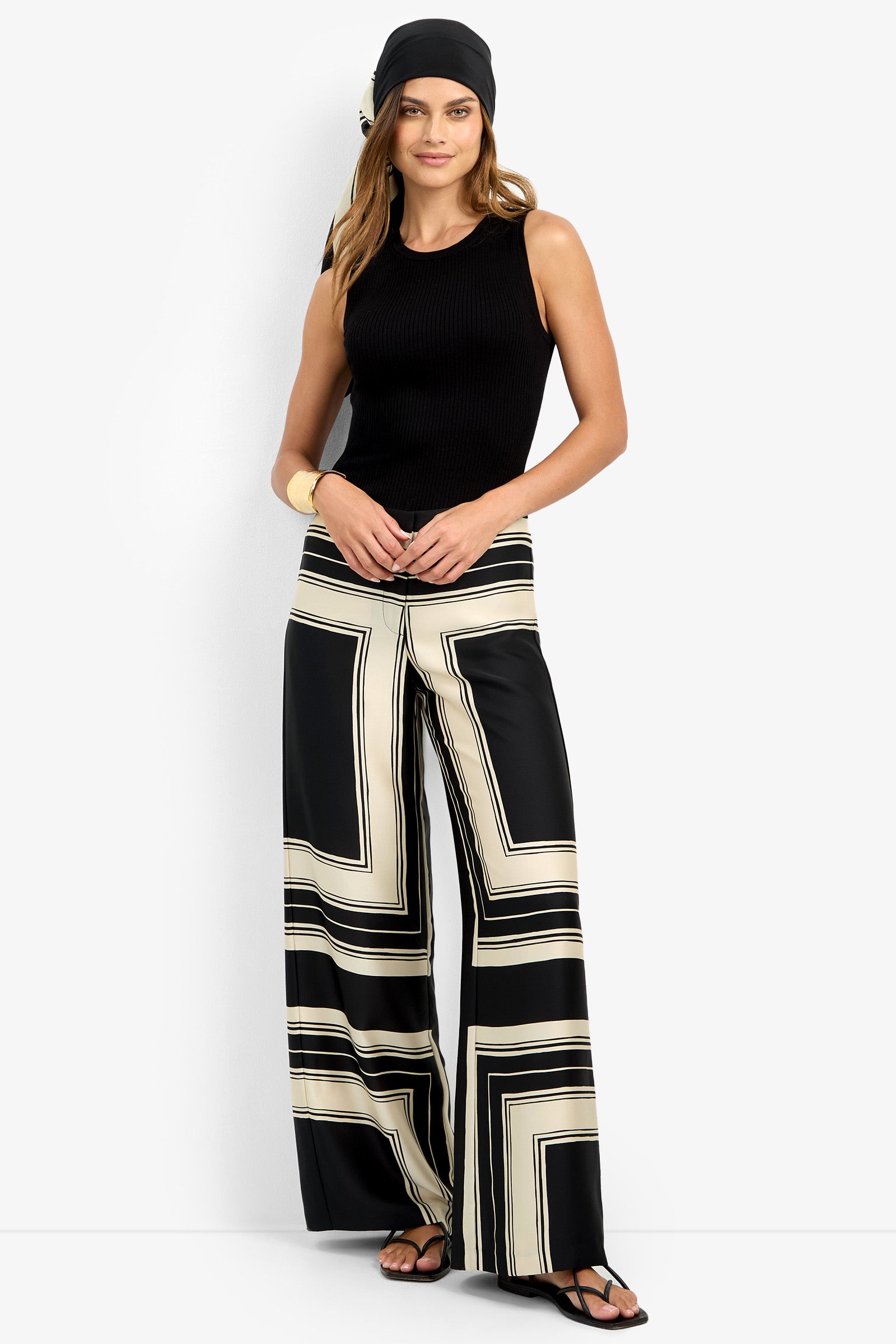 A woman wears the LANAY PANT, featuring wide-leg black and beige geometric print, paired with a black sleeveless top, sandals, headscarf, and gold cuff bracelet against a plain white background.