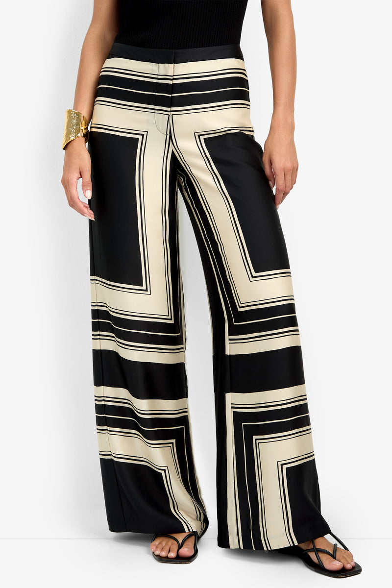 A person wearing the LANAY PANT, featuring a bold black and beige geometric pattern with a wide-leg, high-waisted fit, paired with a black sleeveless top, black sandals, and a large gold cuff bracelet.