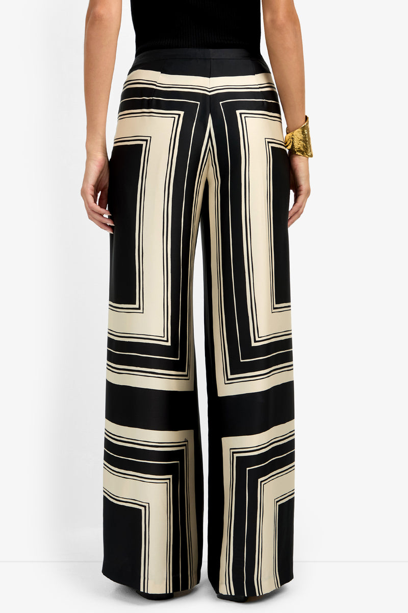A person seen from the back wearing the LANAY PANT, featuring a bold black and cream geometric square print in a high-waisted, wide-leg style, paired with a black top and chunky gold bracelet.