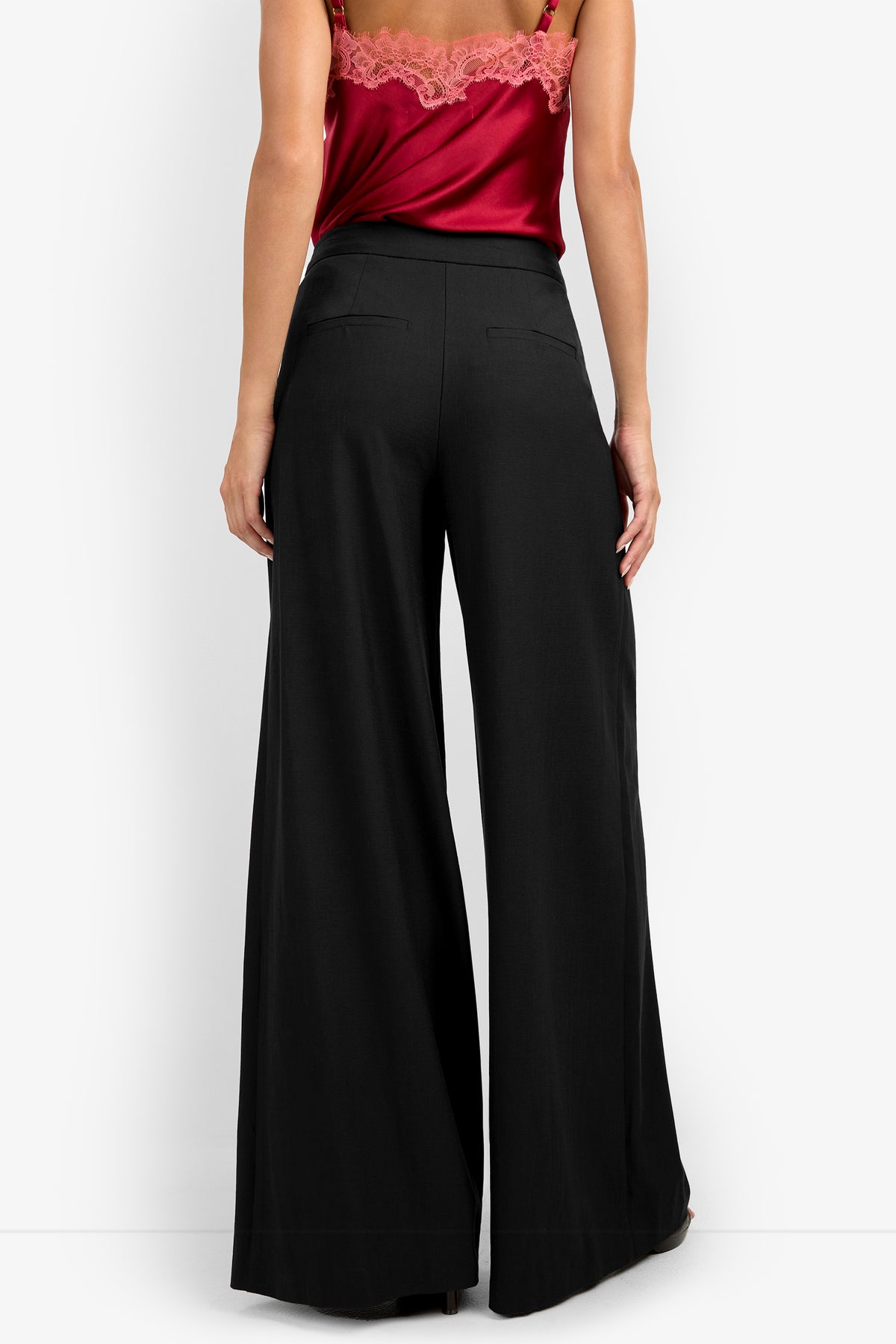 A woman faces away, showcasing the MIMMO PANT—high-waisted, wide-leg black trousers—paired with a red satin lace-trim camisole. The image highlights her lower back and legs.
