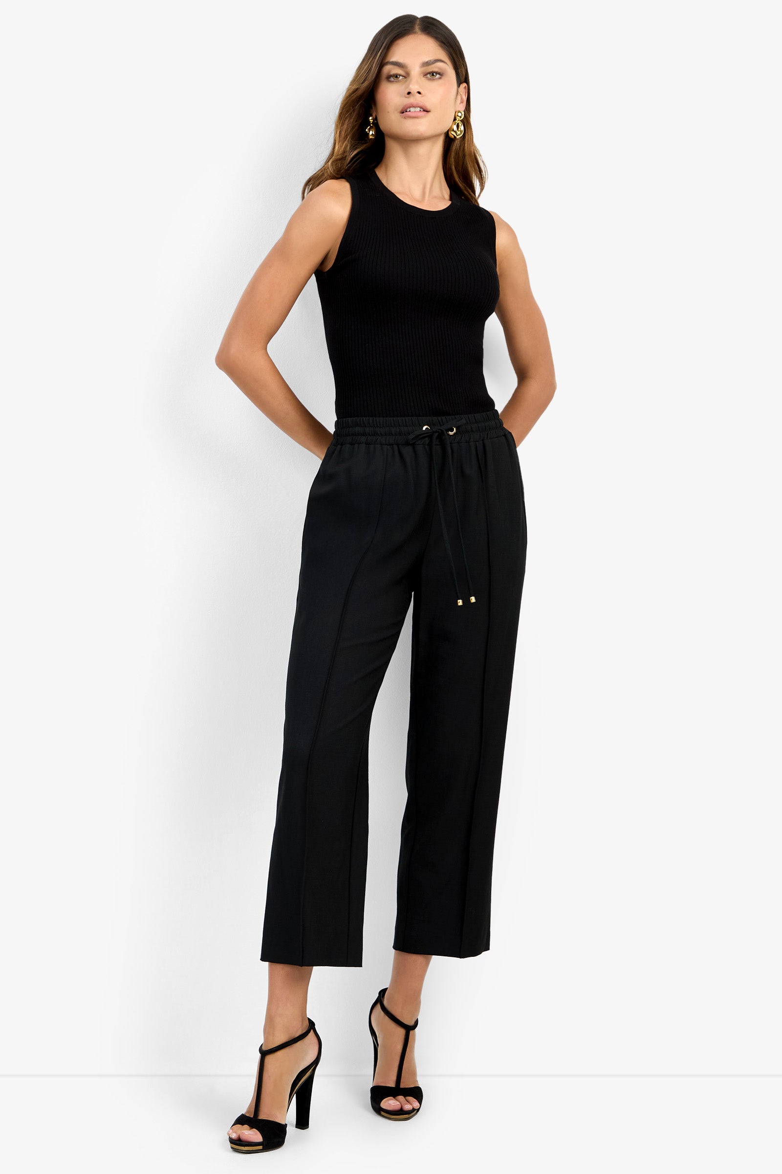 A woman poses against a white backdrop wearing the AVARI PANT—a high-waisted black drawstring pant—paired with a sleeveless black top and black heels, her long hair flowing as she stands with hands in her pockets.