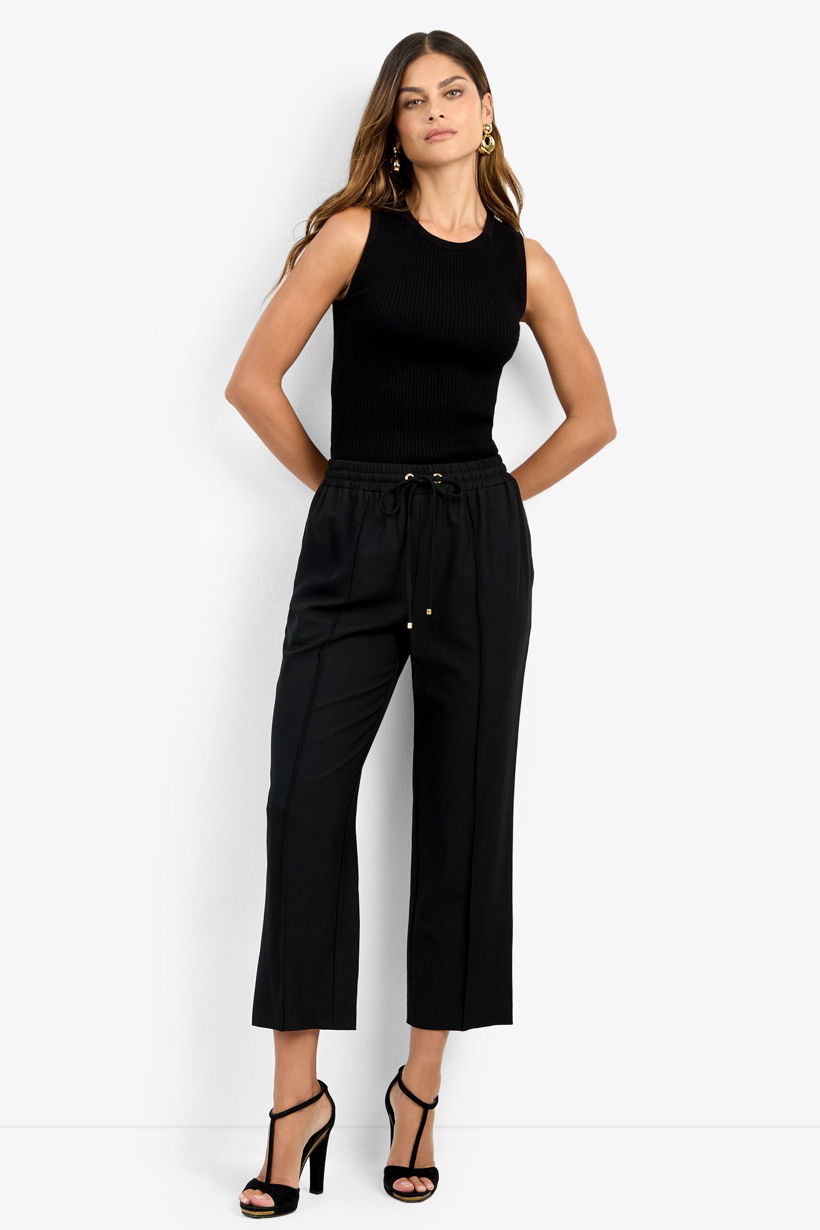 Woman wearing the AVARI PANT—black drawstring trousers—paired with a sleeveless black top and black high-heeled sandals, standing against a plain white background with hands in her pockets.