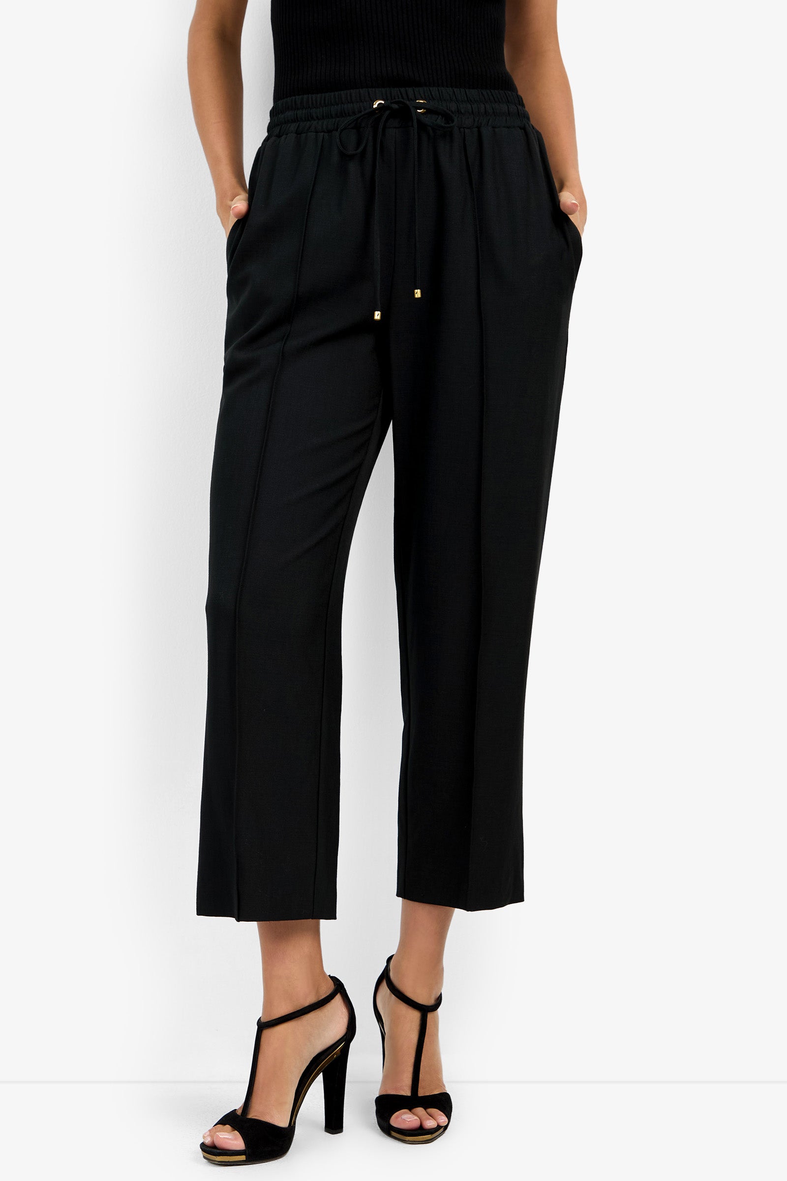 A person wears AVARI PANT—black cropped, wide-leg trousers with a drawstring waist—paired with a black sleeveless top and black high-heeled sandals, standing hands-in-pockets against a white background.