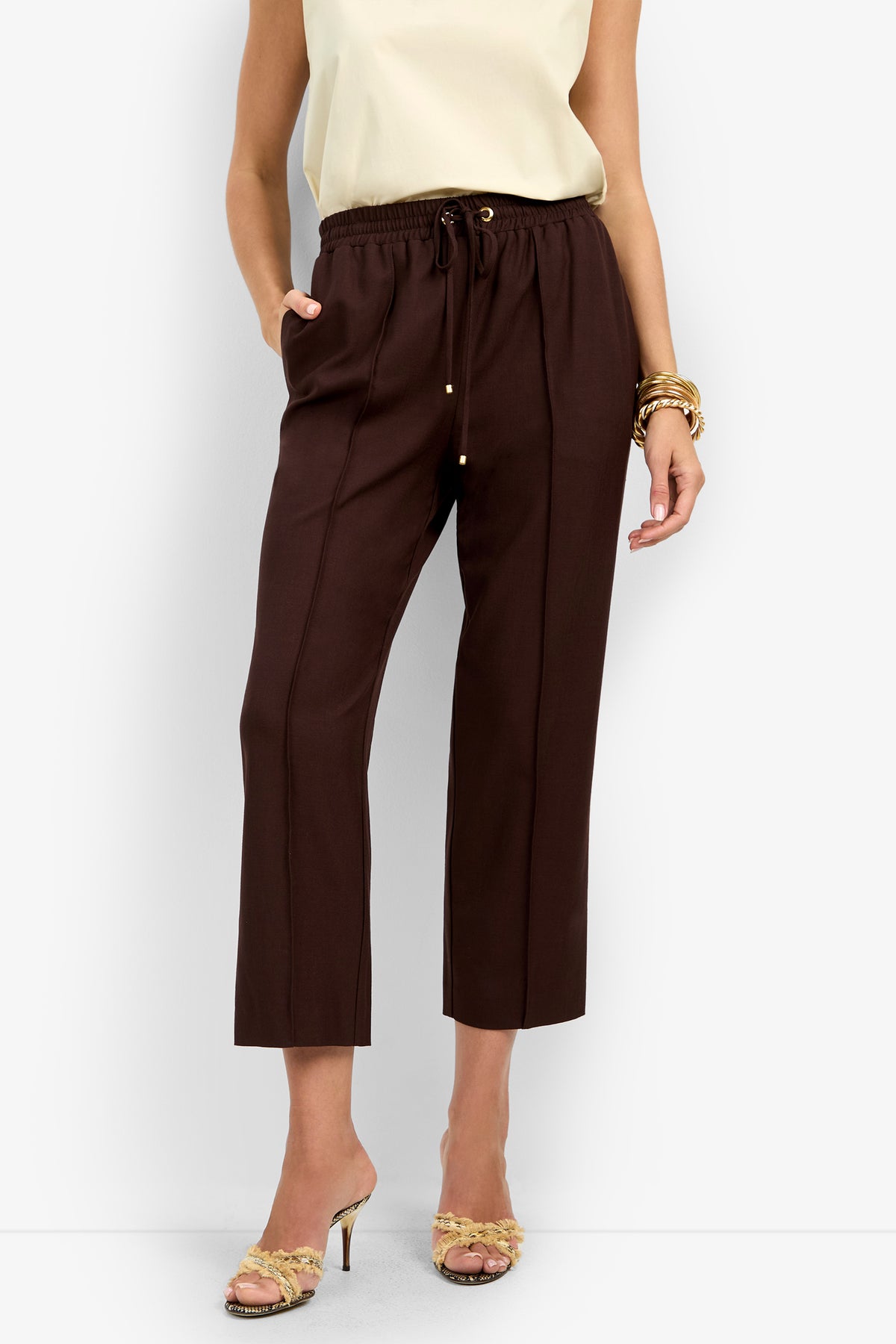 A woman wears the AVARI PANT in dark brown with a sleeveless cream top, gold bracelets, strappy heeled sandals, and stands with one hand in her pocket.