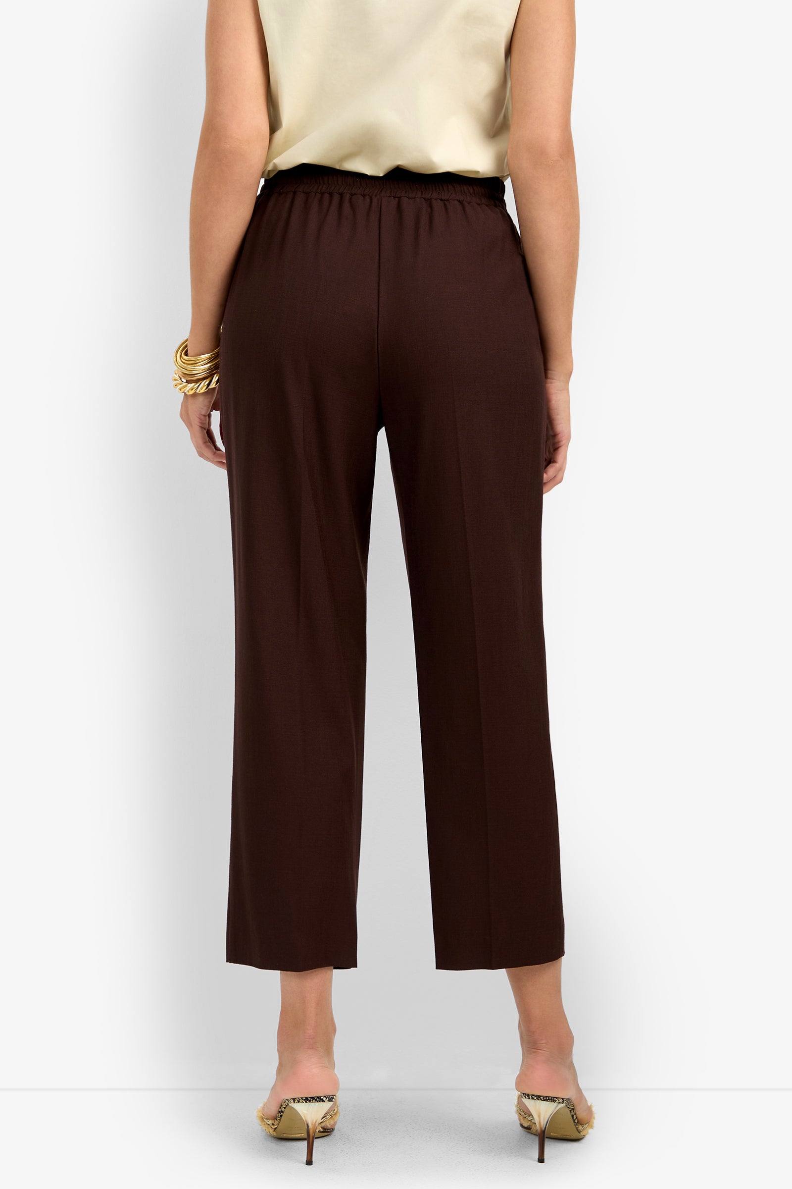 A person stands with their back to the camera wearing a sleeveless beige top, animal print high heels, and AVARI PANT—dark brown wide-leg pants with an elastic waistband.