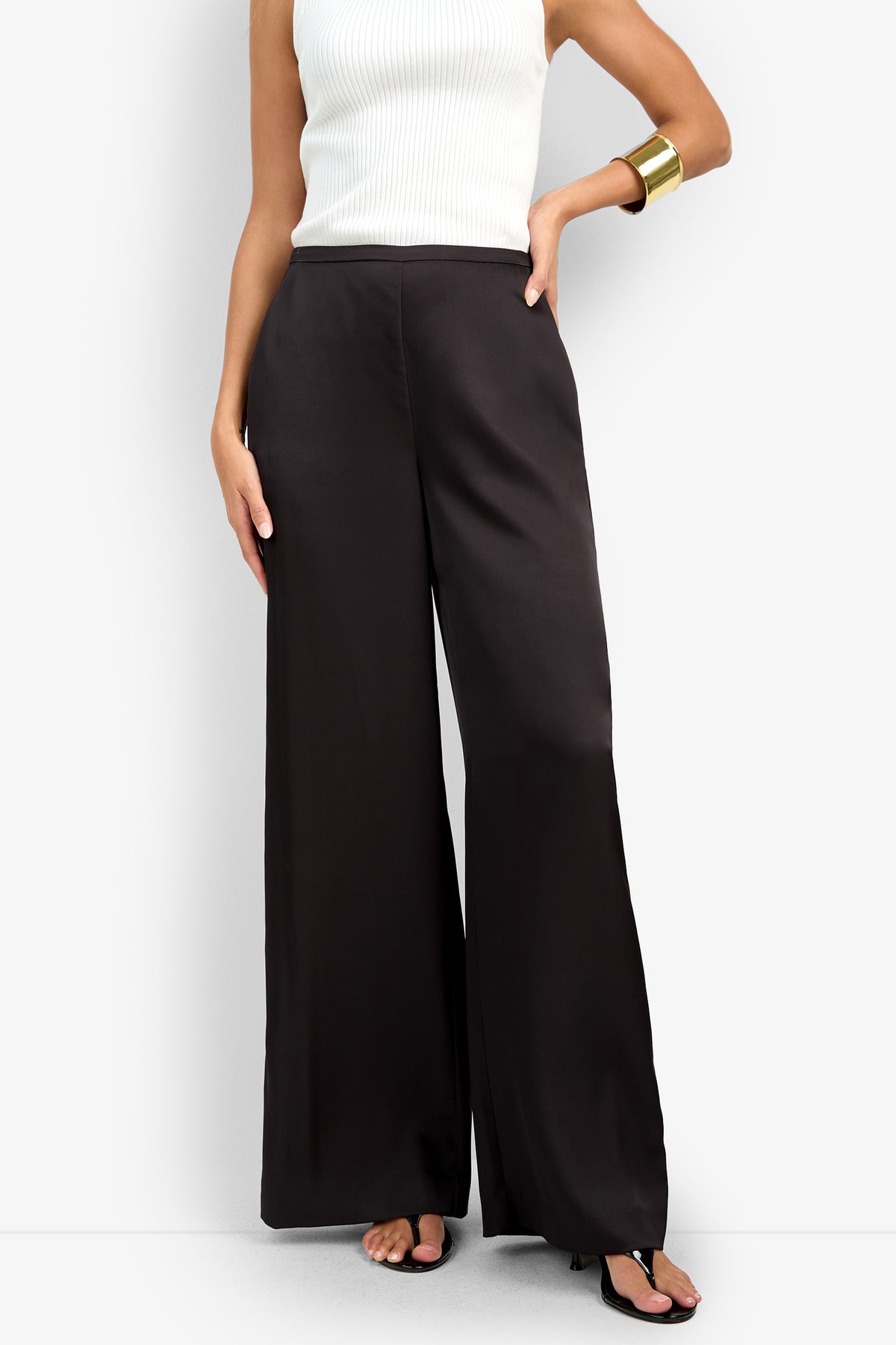 A person wearing the TAVIA PANT—high-waisted, wide-leg black pants—paired with a white sleeveless ribbed top, black heeled sandals, and a gold cuff bracelet, stands against a plain white background.