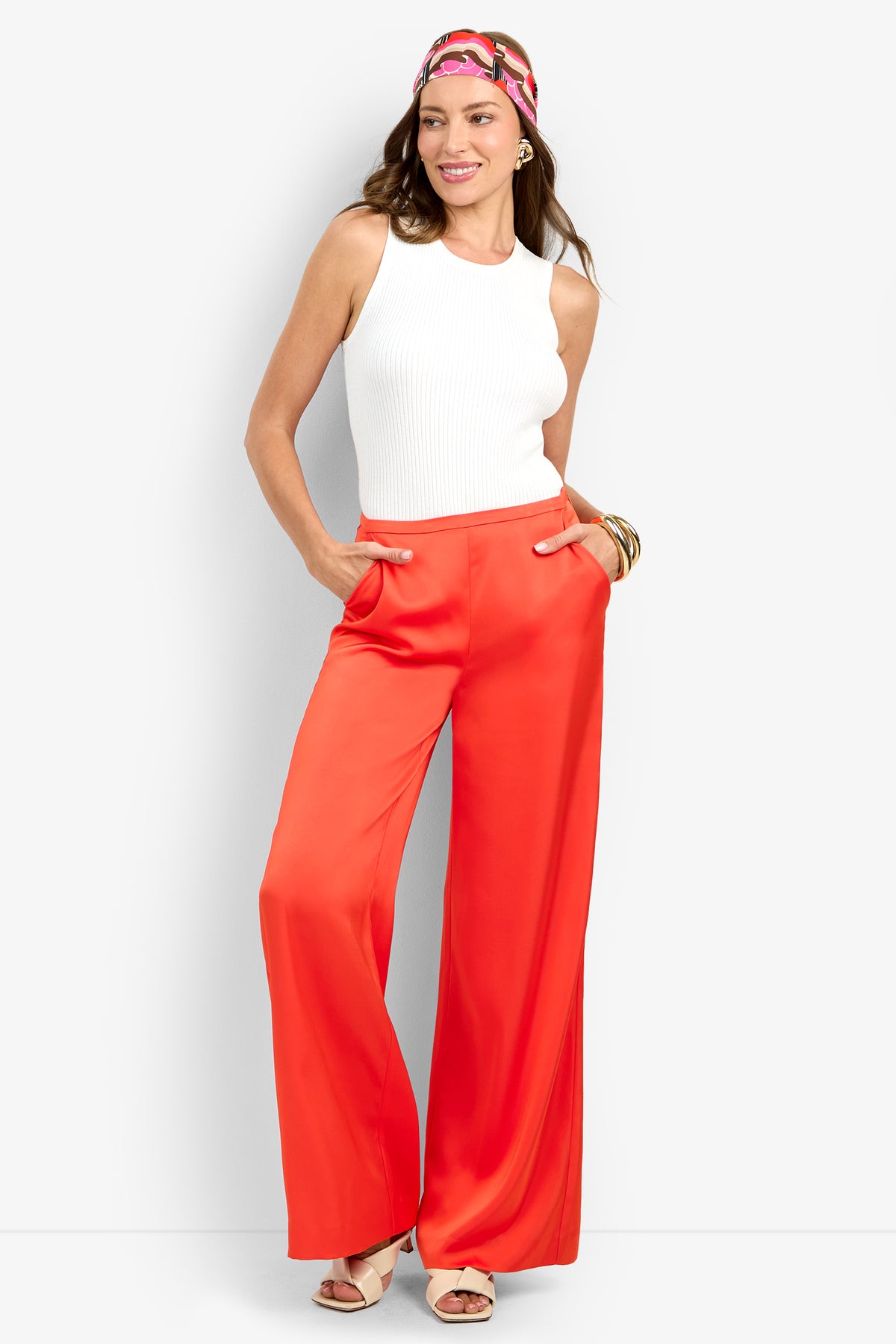 A woman wearing the bright orange TAVIA PANT, a white sleeveless top, beige heels, gold bangles, and a colorful headscarf stands with hands in her pockets, smiling against a plain white background.
