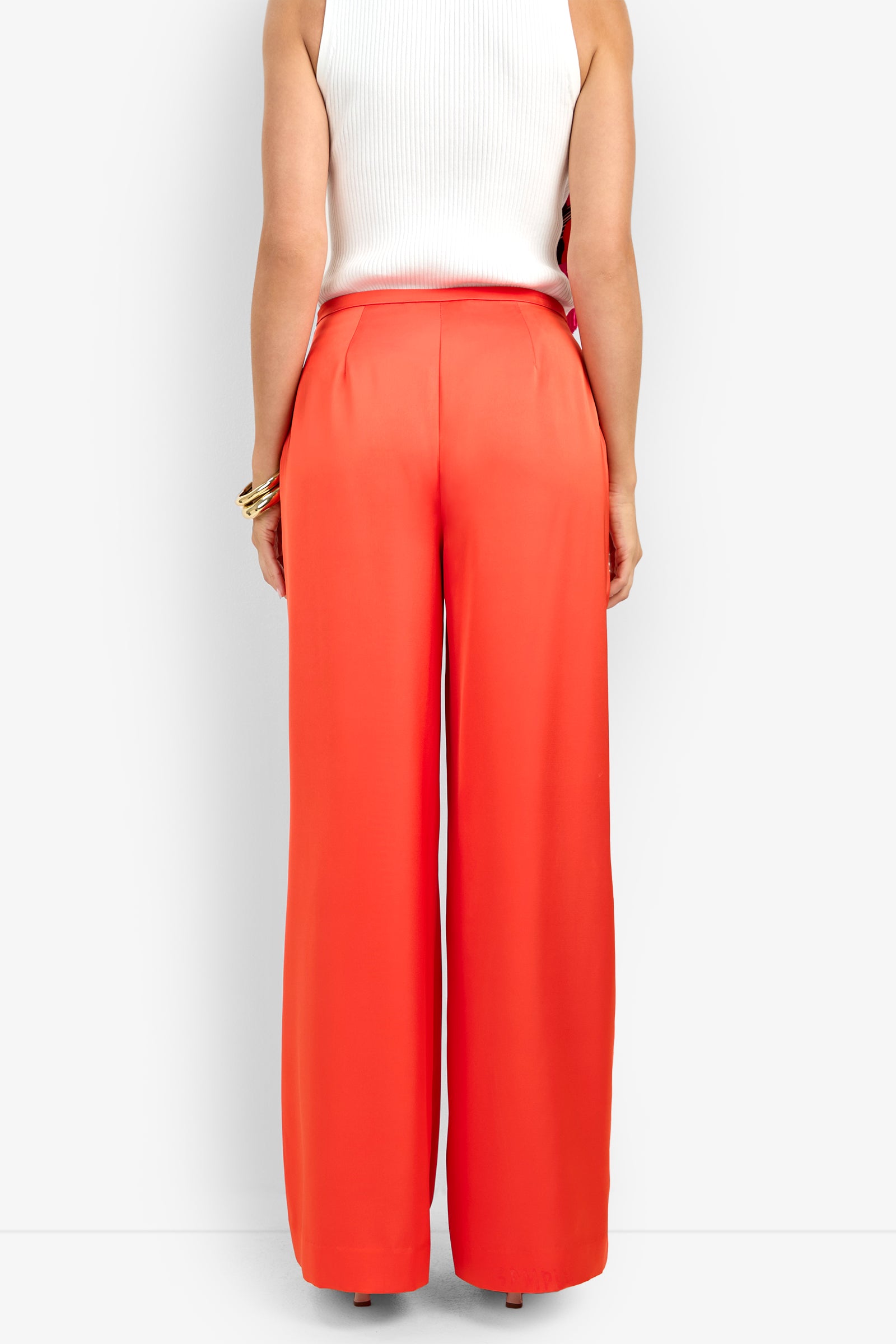 A person is shown from the back wearing the TAVIA PANT in bright orange with a white sleeveless top, standing against a plain white background.