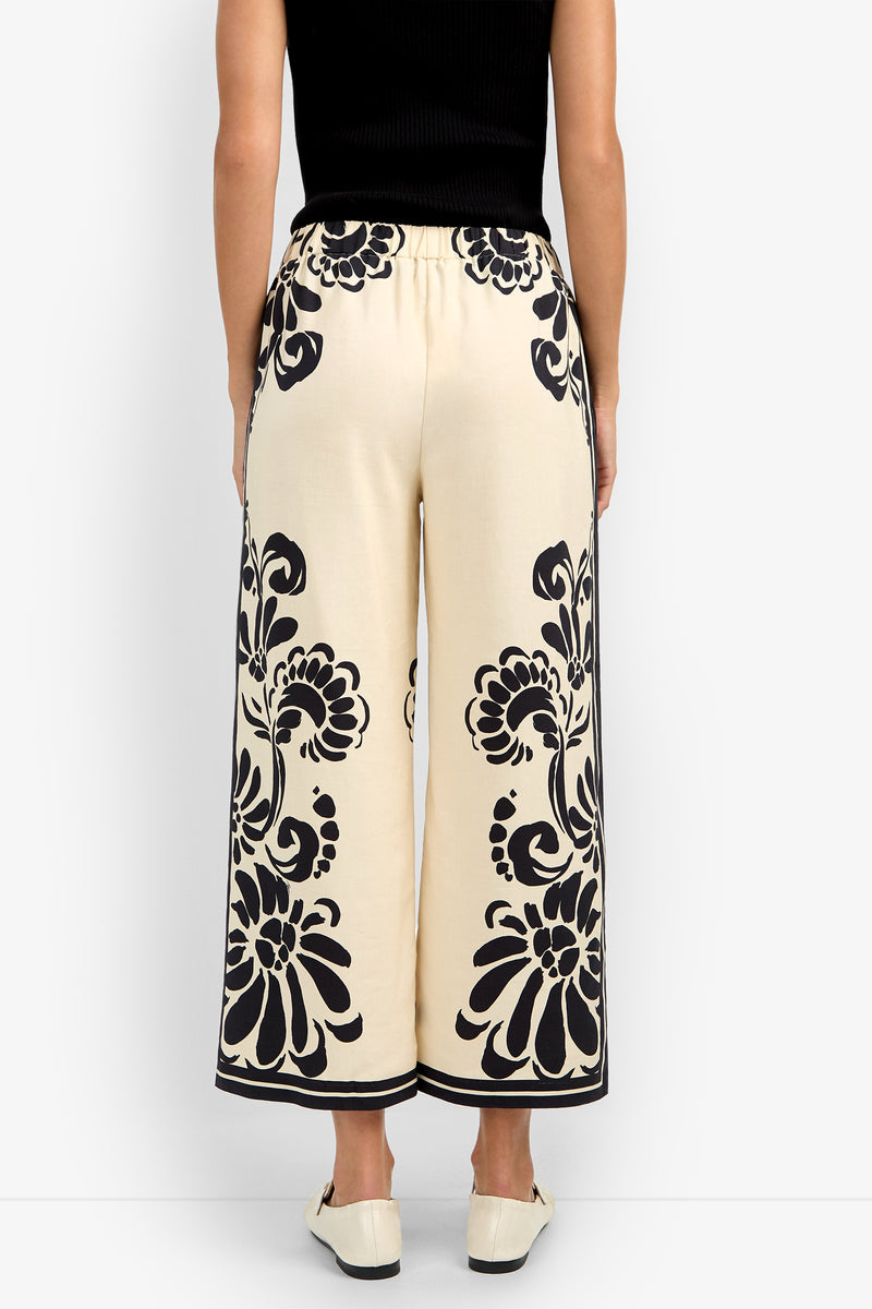 A person shown from the back wears the ESTELLA 2 PANT—cream-colored wide-leg pants with black floral patterns along the sides and hem—paired with white shoes and a black sleeveless top.