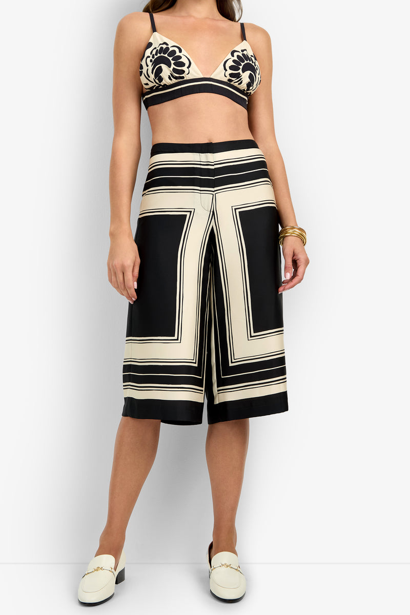 A woman wears the LANAY CROP PANT—cream and black patterned wide-leg culottes—with a matching bralette, white loafers, and gold bracelets, posed against a plain white background.