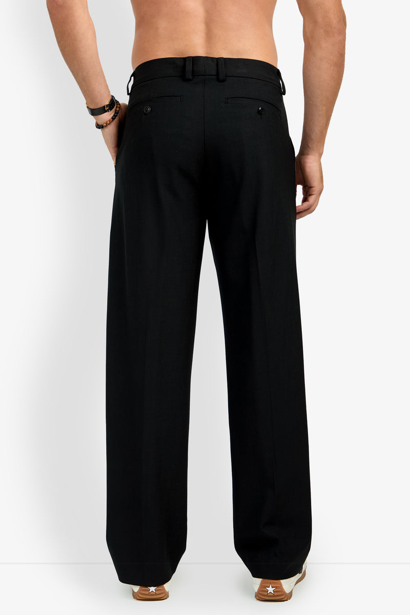 A shirtless person stands facing away, wearing loose-fitting BUCKINGHAM TROUSER in black and brown sneakers with white accents, against a plain white background.