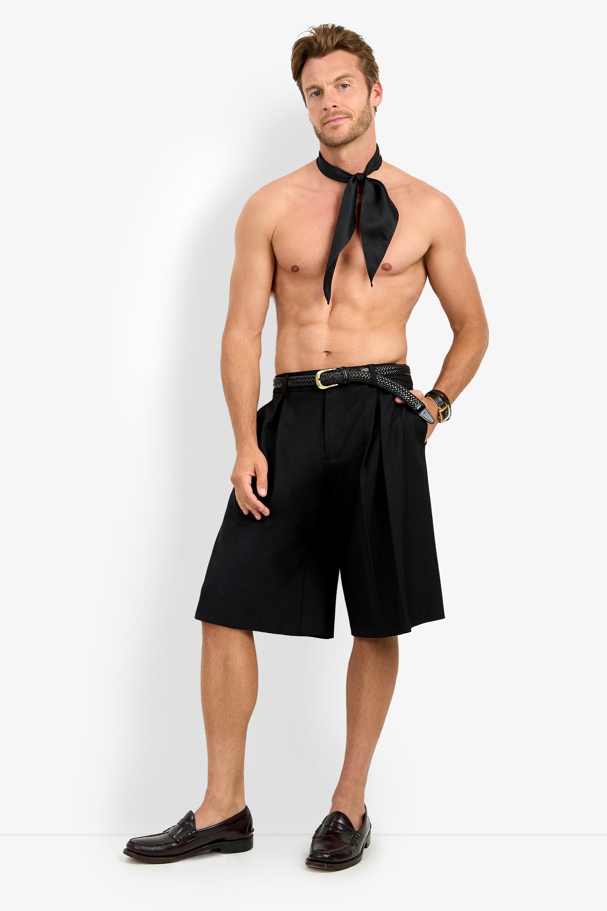 A shirtless man pairs the GALFANO WIDE LEG SHORT with a black necktie, black belt, brown loafers, and bracelets, posing confidently against a plain white background.