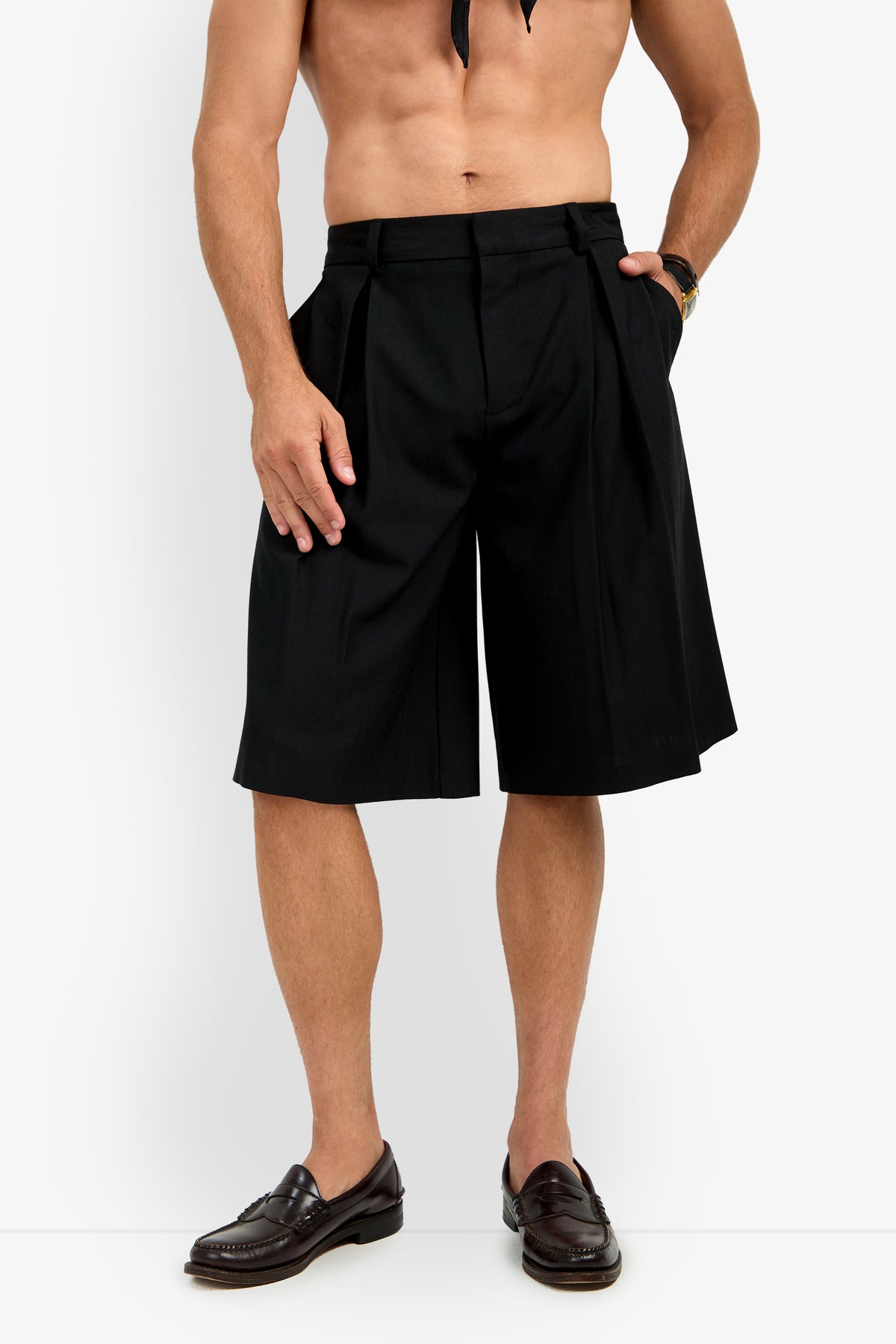 A shirtless man models the GALFANO WIDE LEG SHORT—black, pleated, knee-length shorts—paired with dark loafers against a white background. His hands rest at his sides, and his face is not visible.