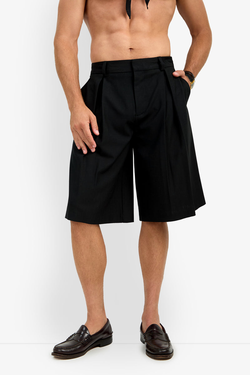 A shirtless man models the GALFANO WIDE LEG SHORT—black, pleated, knee-length shorts—paired with dark loafers against a white background. His hands rest at his sides, and his face is not visible.
